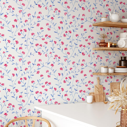 a kitchen with a white table and shelves displaying various kitchen items, including a vase of flowers, a teapot, and a wooden cutting board. The wall behind the table has a floral pattern in shades of blue and red.