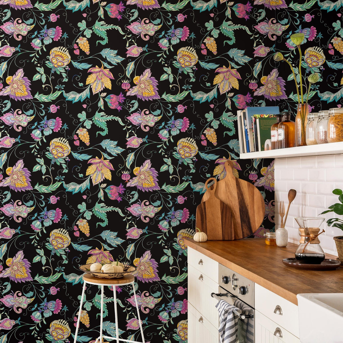 a kitchen with a black and white floral wallpaper, a wooden cutting board, a white shelf with various items, and a white table with a bowl of fruit.