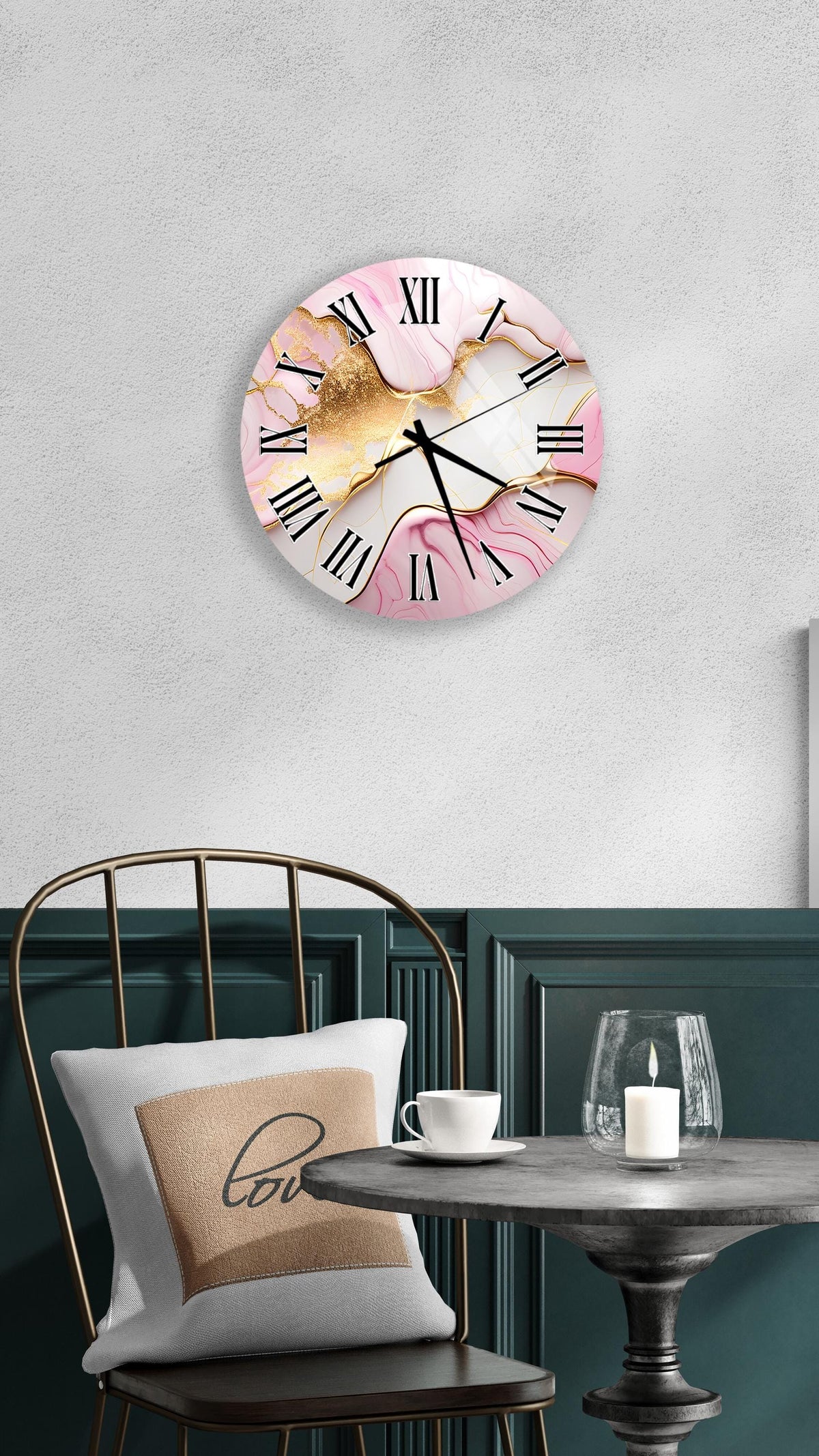 a wall clock with a pink and gold marble design, mounted on a wall above a table with a white tablecloth. The table has a glass candle and a white coffee cup on it. A white pillow with the word "love" printed on it is also present on the table.