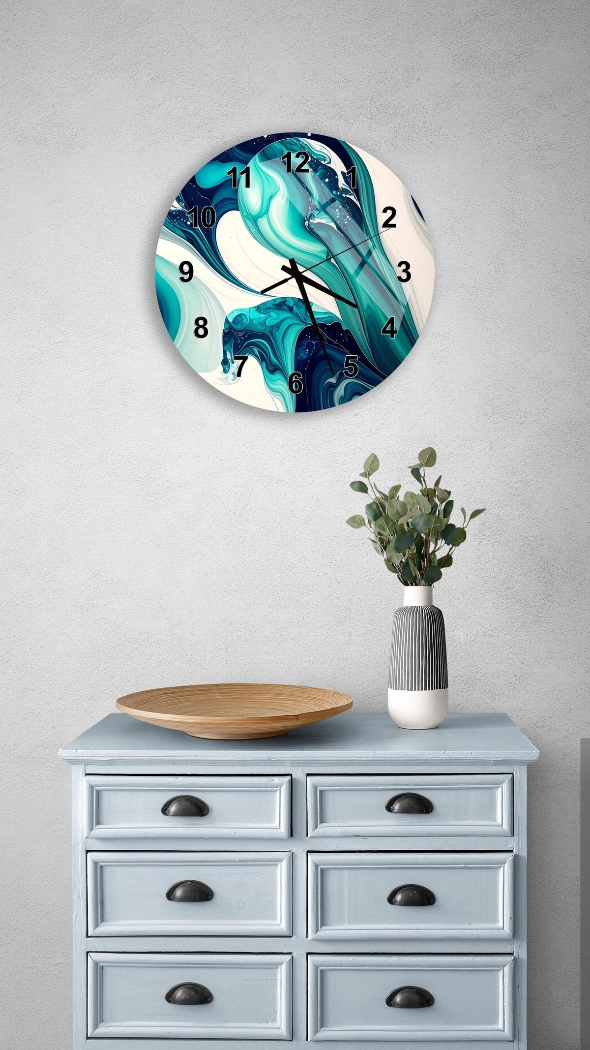 a wall clock with a blue and white abstract design, mounted on a wall above a light blue dresser with six drawers. The dresser also has a vase with greenery and a wooden bowl on top.