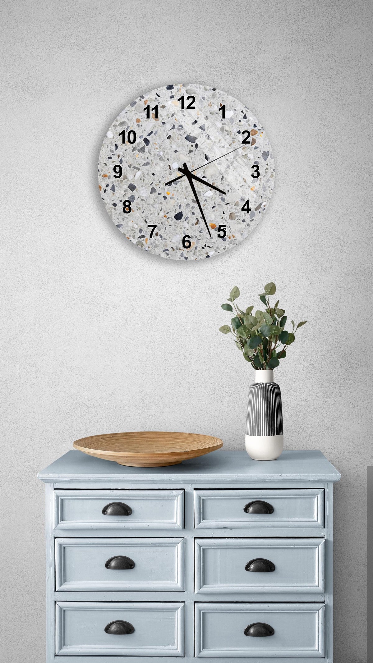 a wall clock with a speckled design, mounted on a wall above a light blue wooden dresser with six drawers. The dresser also has a vase with greenery on it, and a wooden plate on top.