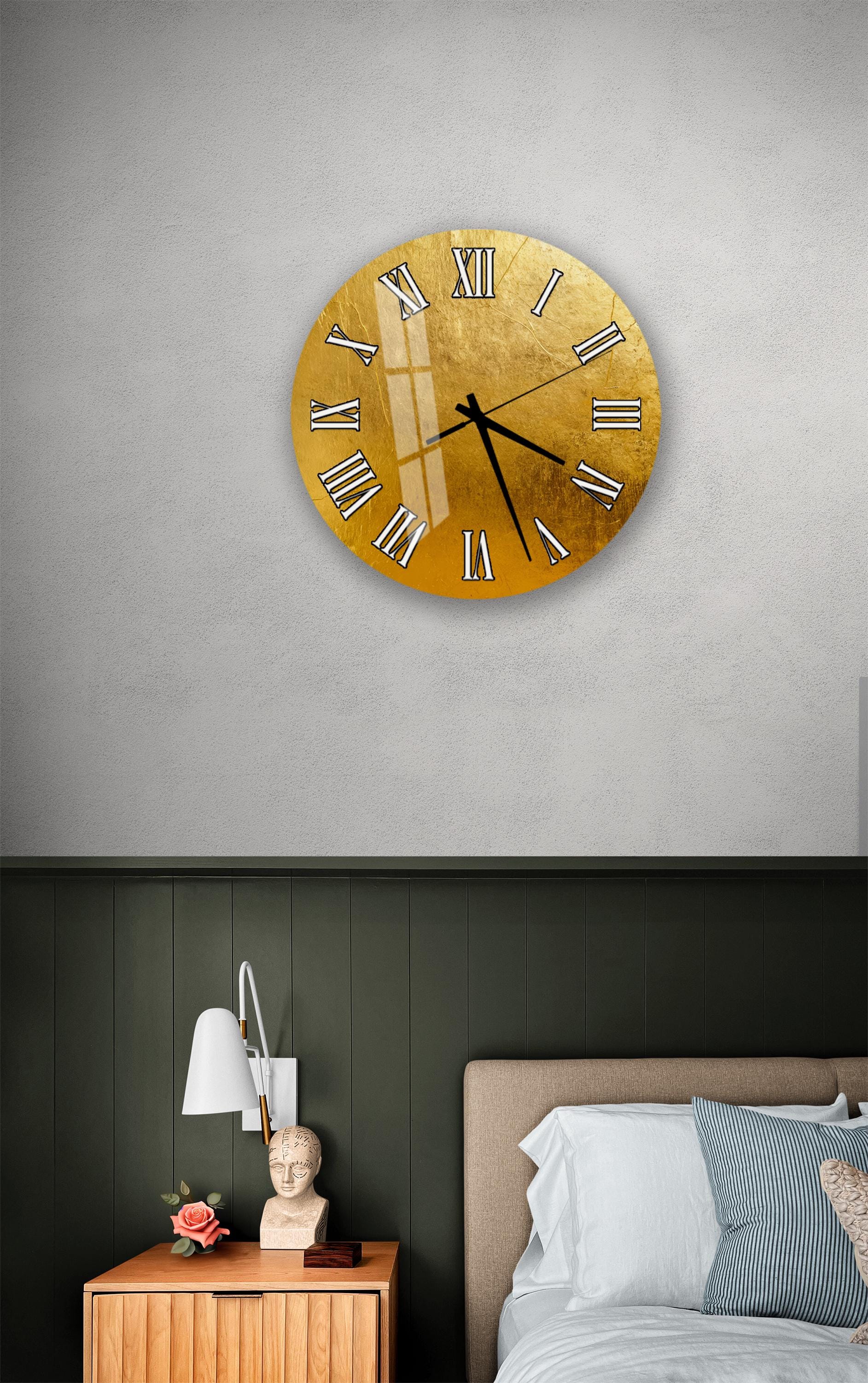 a bedroom setting with a large gold-colored clock on the wall above a bed. The clock has Roman numerals and appears to be a wall-mounted design. The room has a green wall, a wooden nightstand with a lamp, and a bed with white sheets.