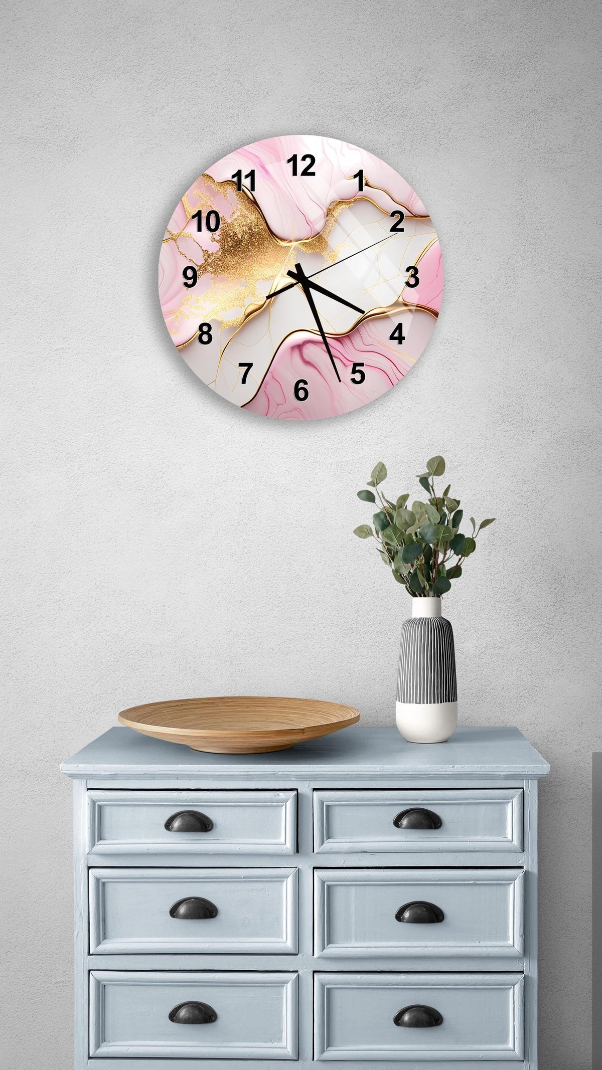a wall clock with a pink and white marble design, mounted on a wall above a light blue dresser with six drawers. The dresser also has a vase with greenery on it, and there is a wooden plate on the dresser.