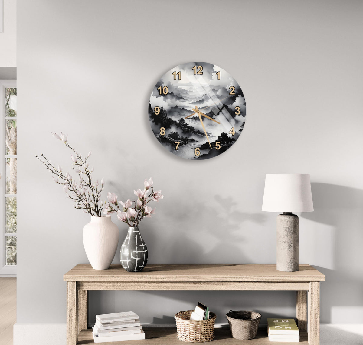 a wall clock with a black and white landscape design, a wooden table with a lamp, a vase with pink flowers, and a stack of books.
