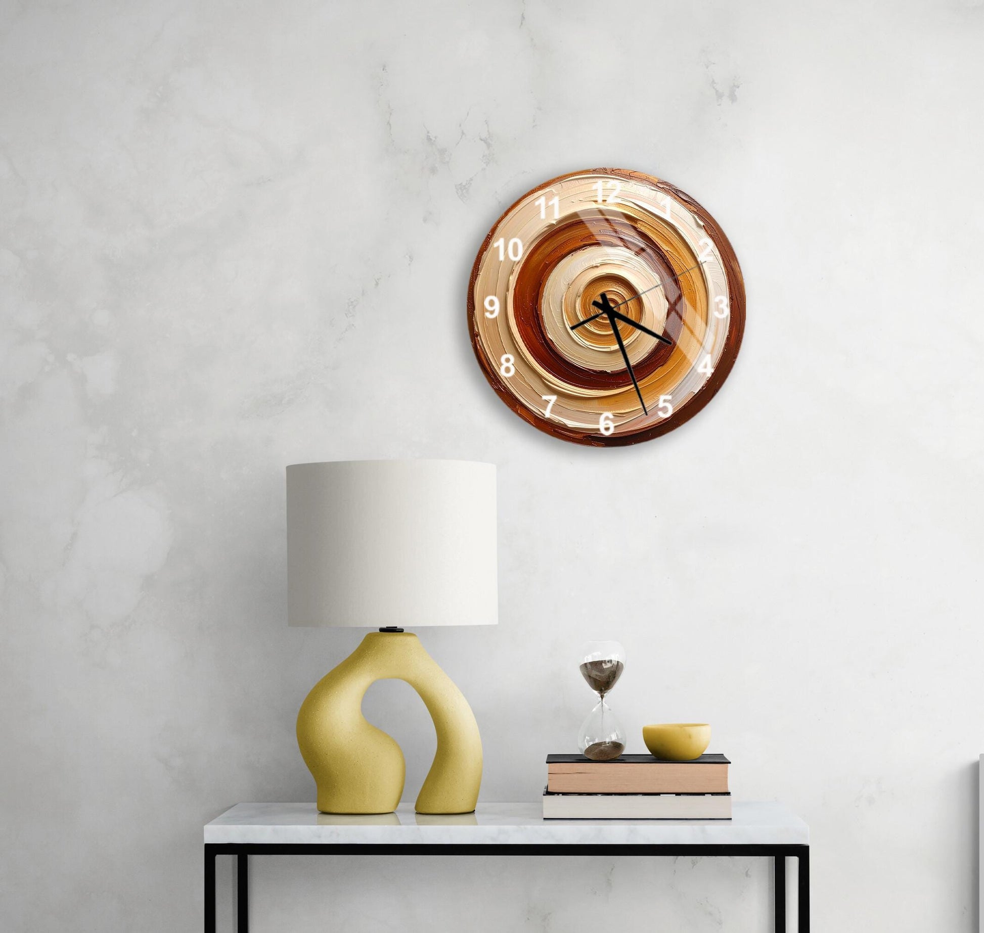 a modern wall clock with a brown and orange design, mounted on a white wall above a white table with a yellow lamp and a stack of books.