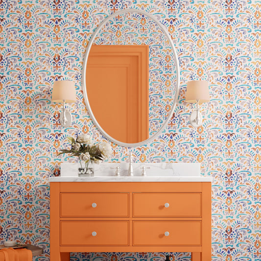 a bathroom with a vibrant orange vanity and a large mirror above it. The vanity has a white countertop and two lamps on either side. The walls are covered in a colorful, patterned wallpaper, and there is a vase of flowers on the vanity.