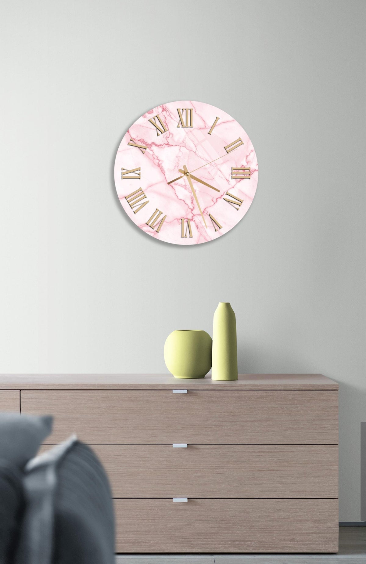 a pink marble clock with Roman numerals on a wall, above a wooden dresser with three drawers.