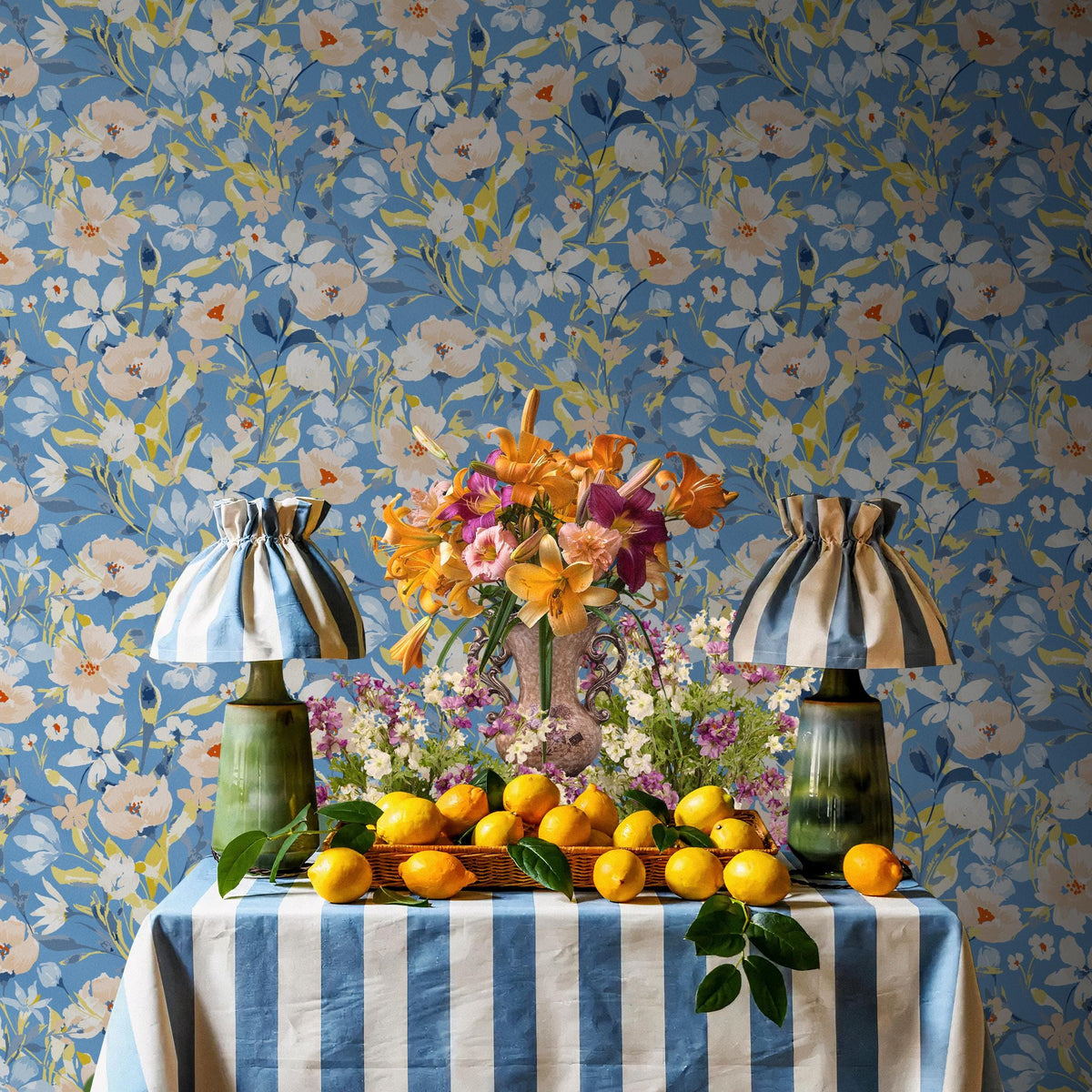 a table with a blue and white striped tablecloth, a vase of flowers, and a basket of lemons. The table is set against a blue floral wallpaper.