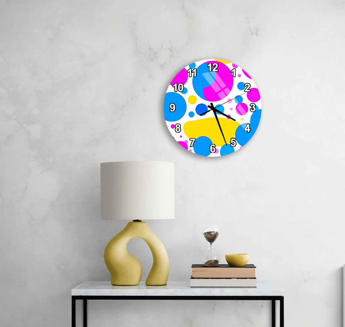 a colorful abstract clock with a white base and a vibrant design, hanging on a wall above a white table with a yellow lamp and a stack of books.