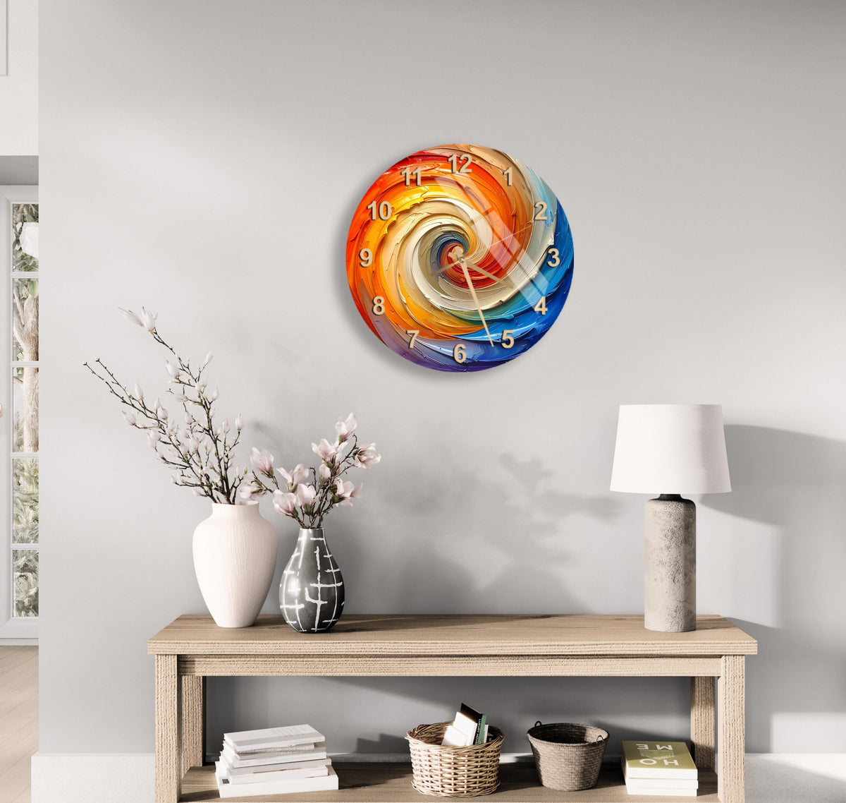 a modern, colorful clock with a spiral design hanging on a wall above a wooden table with various decorative items, including a vase with flowers, a lamp, and a stack of books.