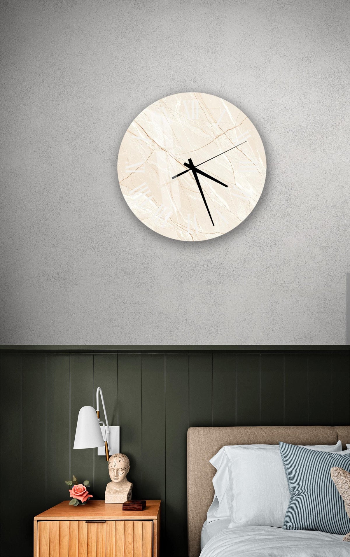 a bedroom setting with a round clock on the wall above a bed. The clock has a marble-like design and is mounted on a gray wall. The bed has a wooden headboard and is covered with white sheets. There is also a nightstand with a lamp and a small statue on it.