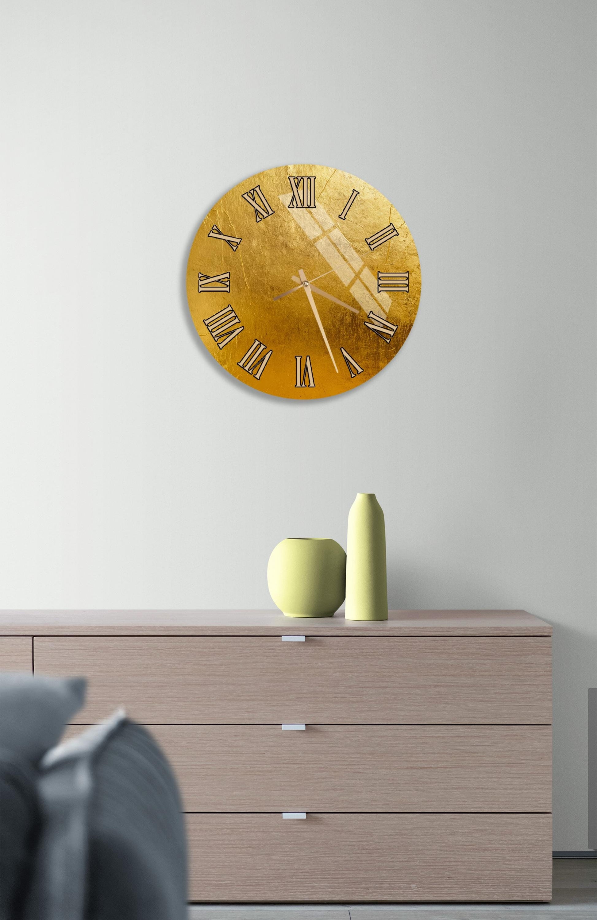 a modern living room with a large gold-colored clock hanging on the wall above a wooden dresser. The dresser has three drawers and a vase on top.