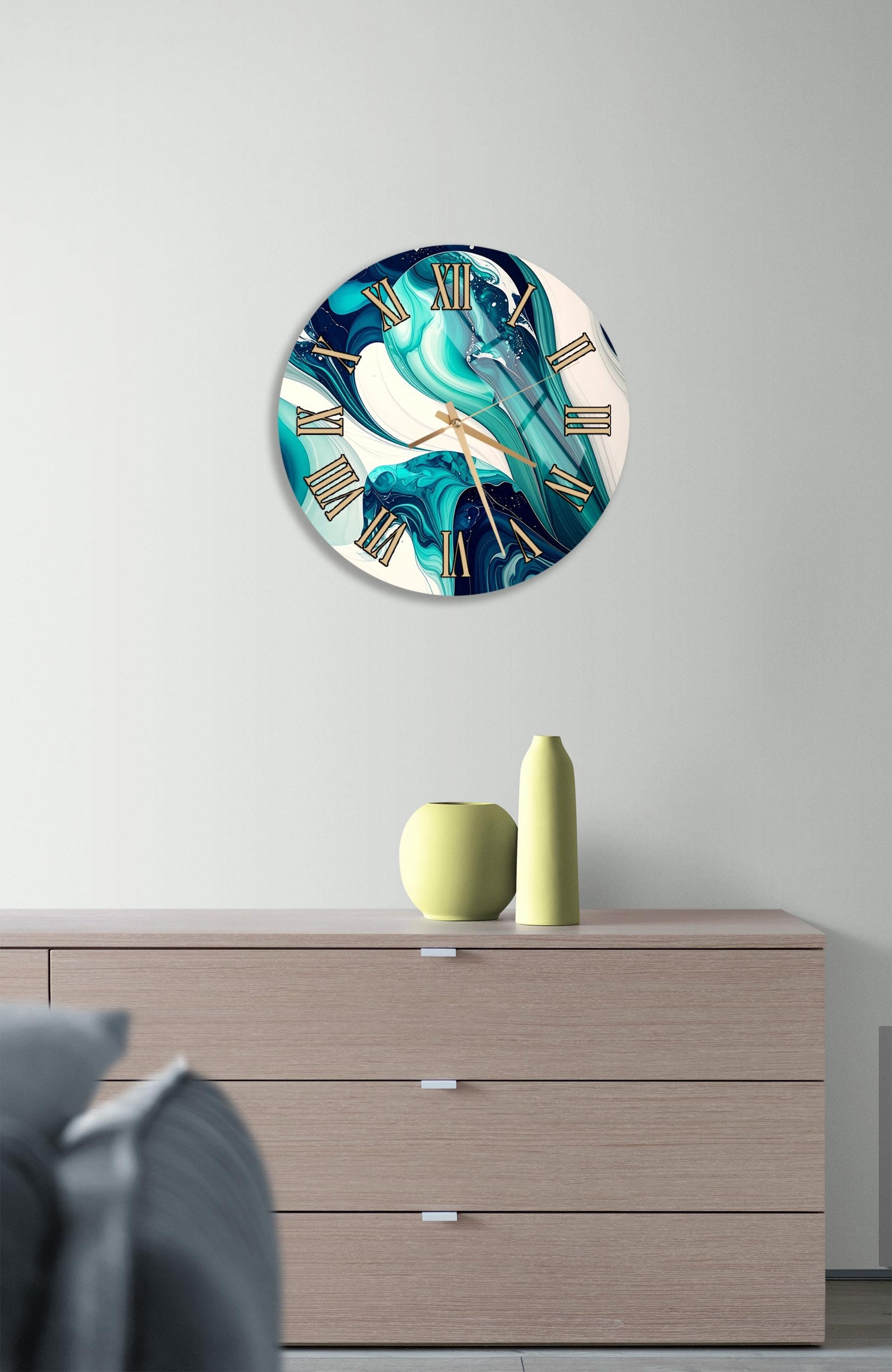 a modern clock with a turquoise and white design, hanging on a wall above a wooden dresser with three drawers. The dresser also has a small vase and a yellow bottle on top of it.