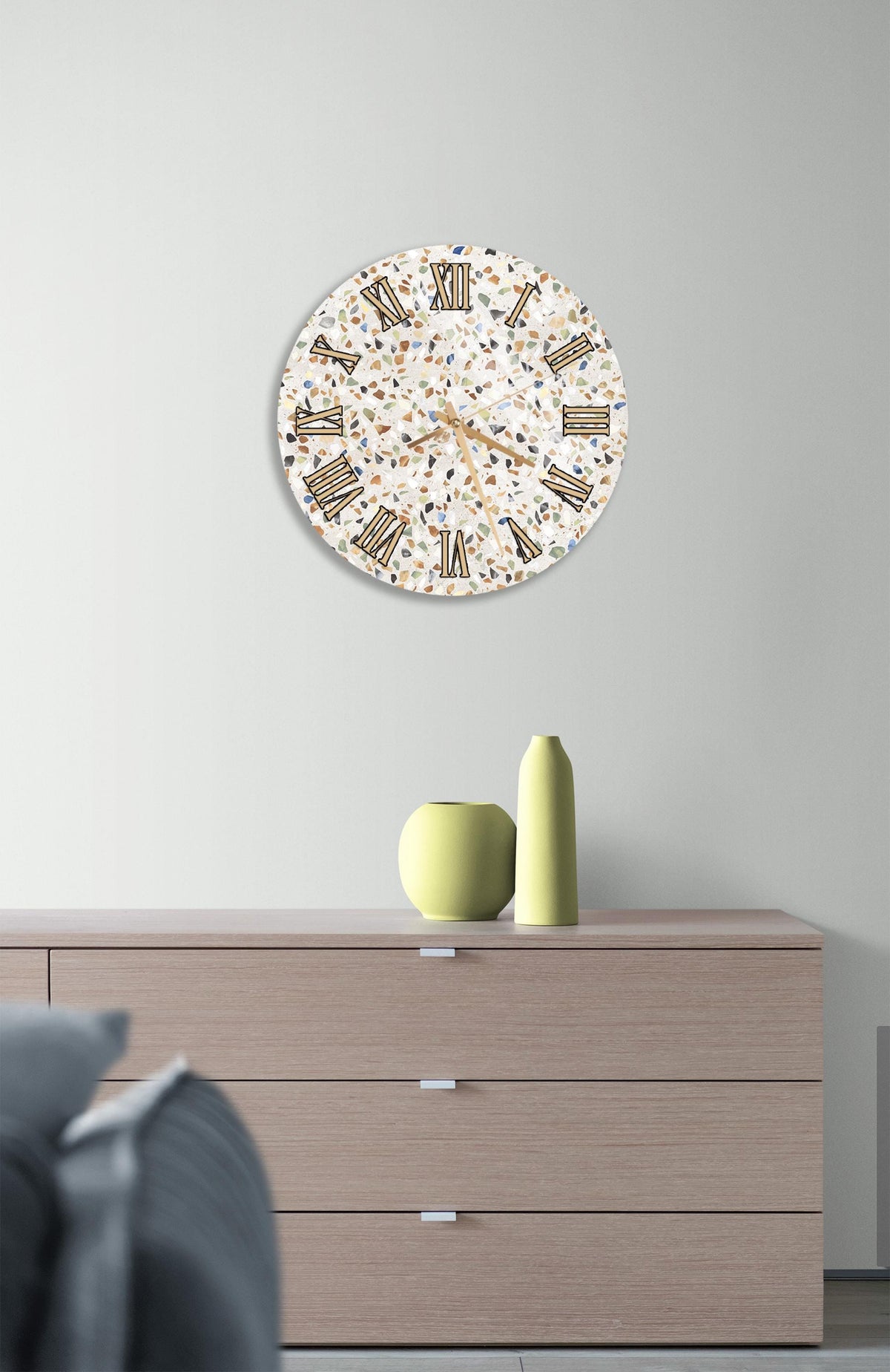 a modern living room with a large, round wall clock displaying Roman numerals. The clock is mounted on a light-colored wall above a wooden dresser with three drawers. On the dresser, there are two small vases and a yellow vase, adding a decorative touch to the room.