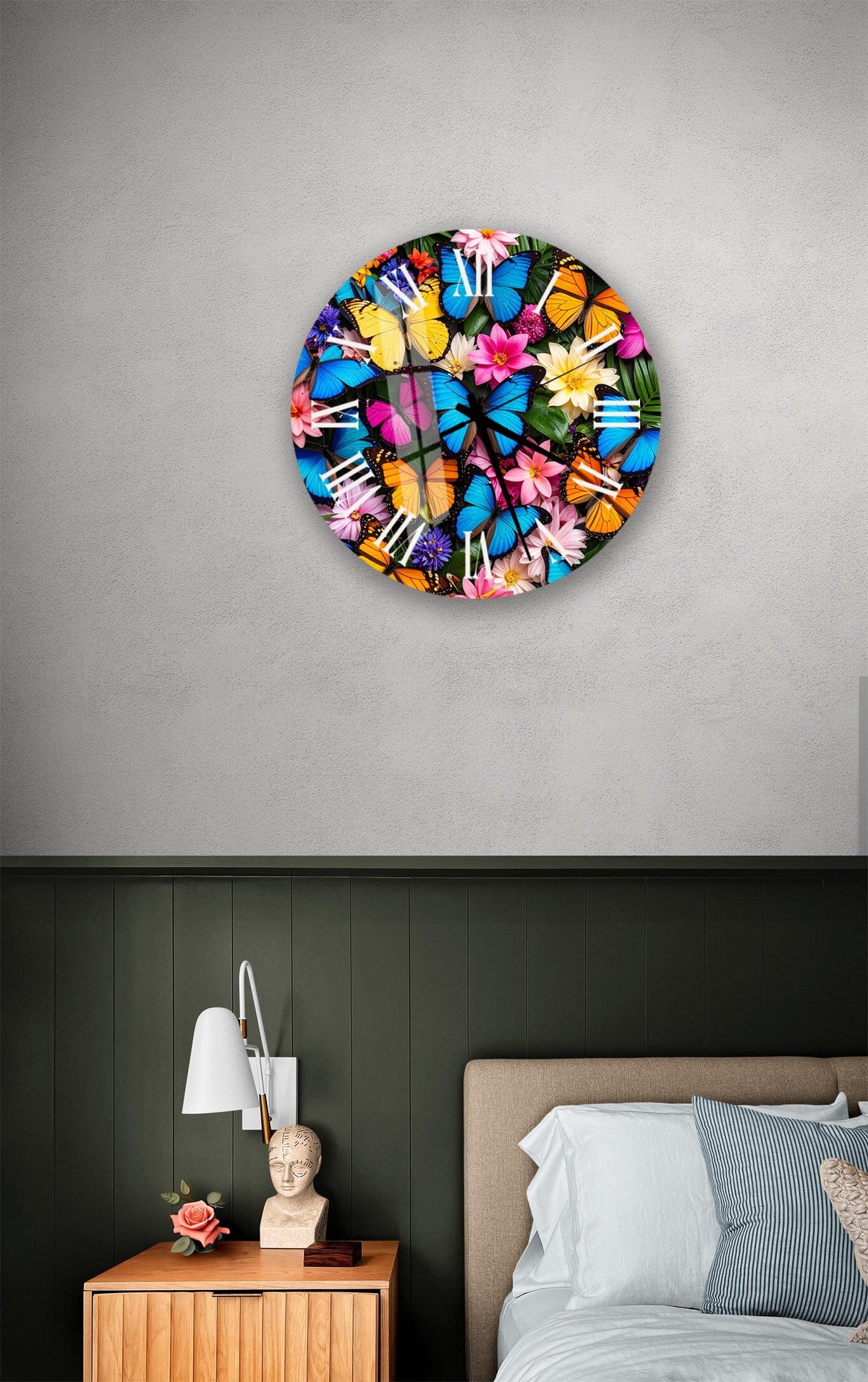 a bedroom setting with a large, colorful clock hanging on the wall above a bed. The clock has a floral design and is surrounded by a variety of vibrant flowers and butterflies. The room also includes a nightstand with a lamp and a statue, creating a cozy and inviting atmosphere.