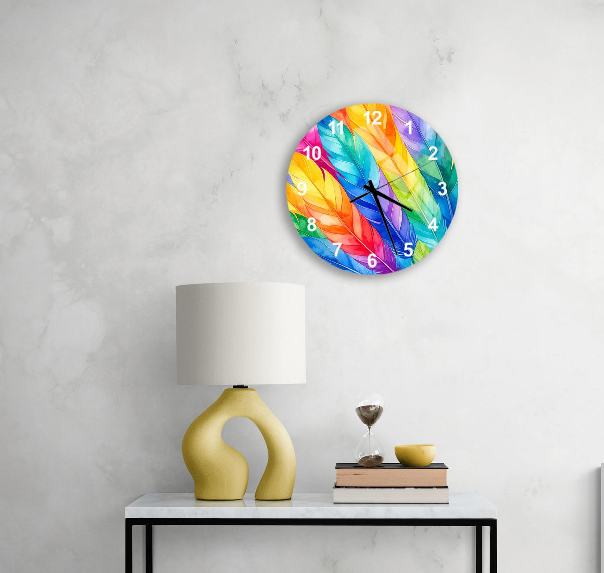 a colorful wall clock with a vibrant feather design, mounted on a wall above a white table with a yellow lamp and a stack of books.