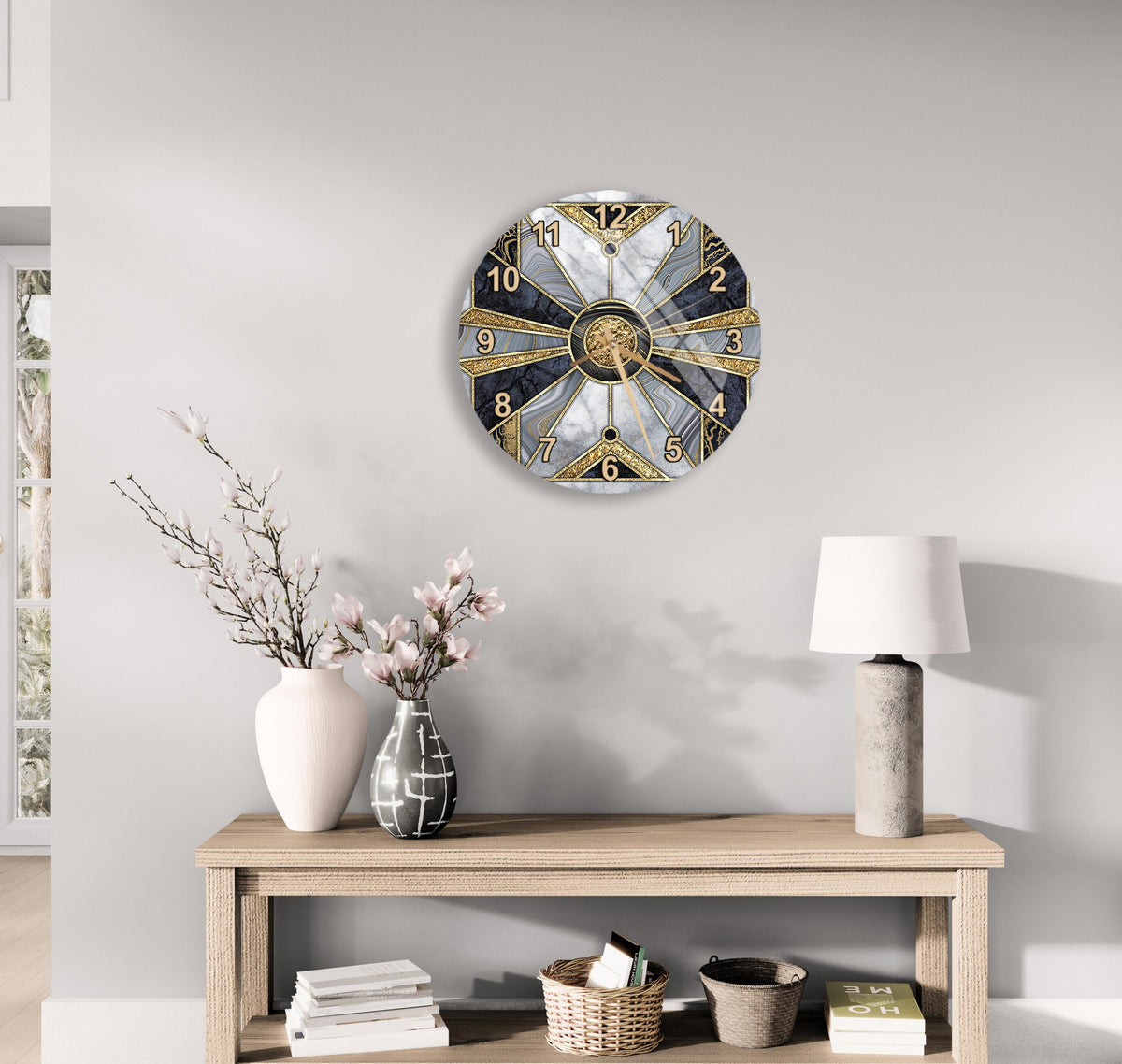 a modern, round clock with a gold and black design, mounted on a wall above a wooden table with a lamp and decorative items.