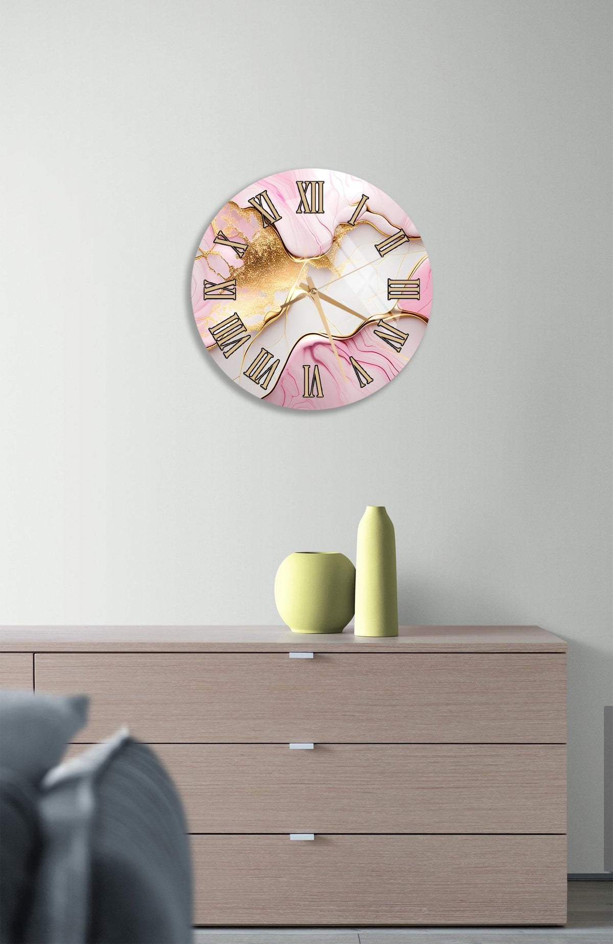 a wall clock with a pink and gold design, mounted on a wall above a wooden dresser with a vase and a small green pot on top.