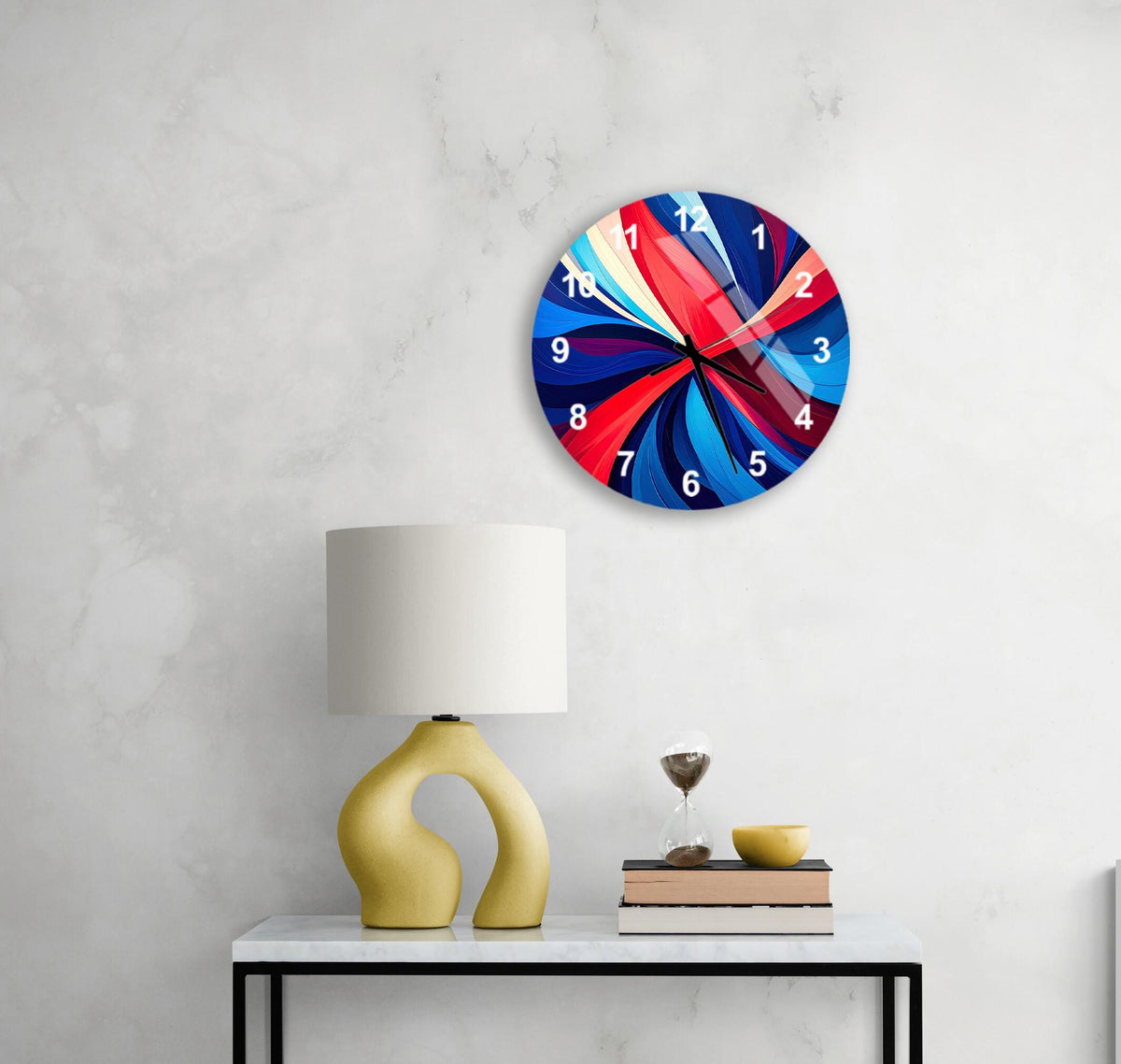 a modern, abstract wall clock with a vibrant design, mounted on a wall above a white table with a yellow lamp and a stack of books.