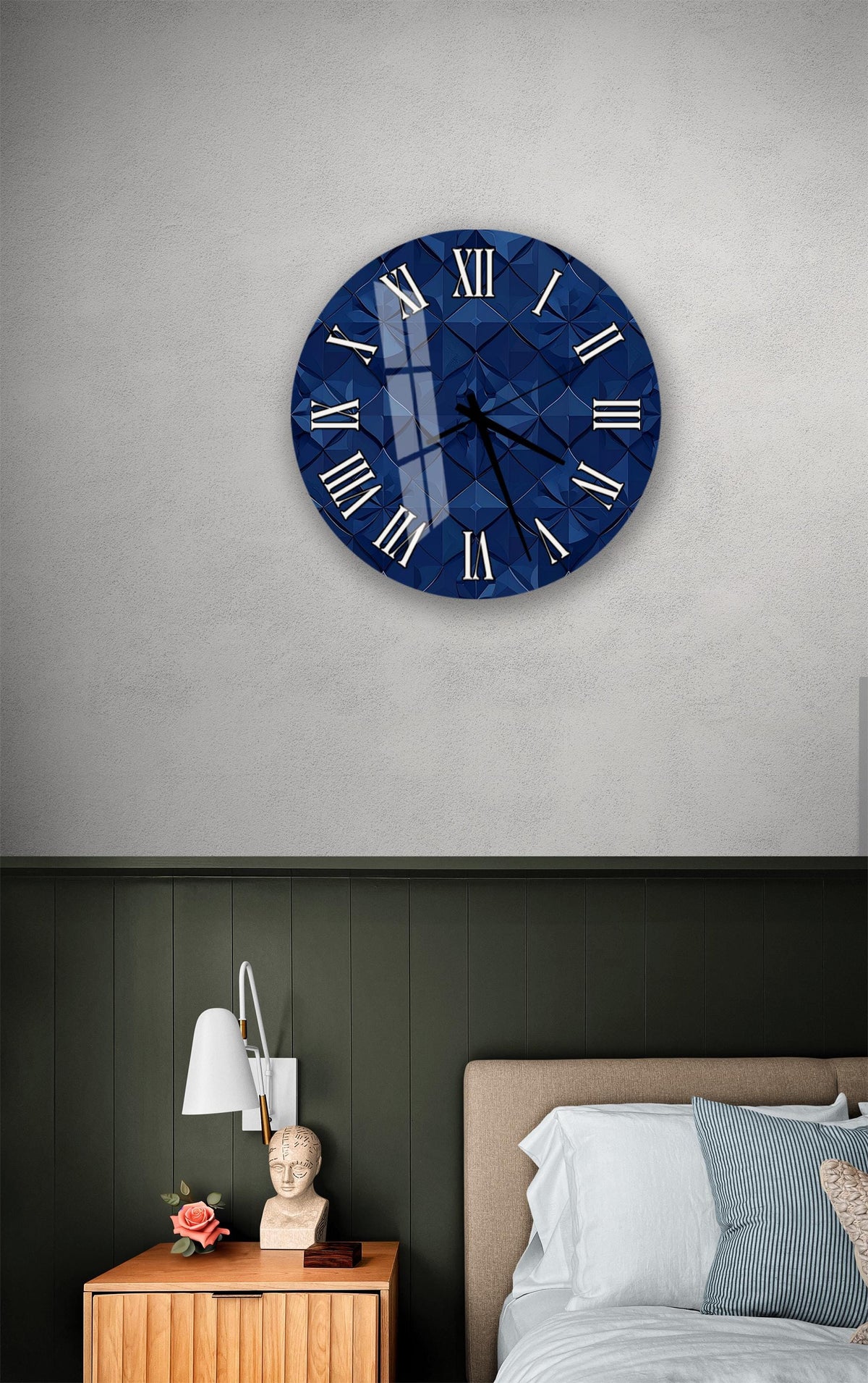 a bedroom setting with a large blue clock on the wall above a bed. The clock has a diamond pattern and Roman numerals. The room has a green wall and a wooden nightstand with a lamp and a vase of flowers.
