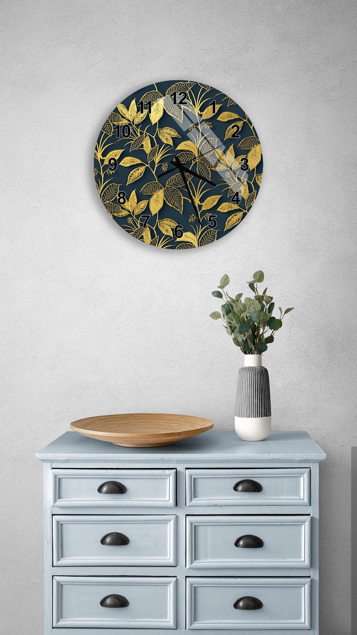 a wall clock with a gold leaf design, a wooden bowl, and a vase with greenery on a light blue dresser.