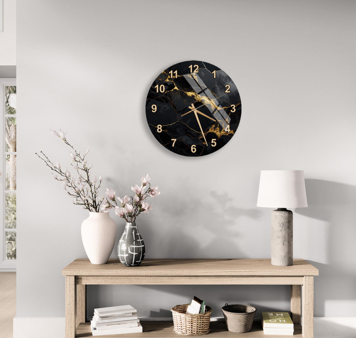 a modern living room with a large black and gold wall clock hanging above a wooden table. The table has a lamp, a vase with flowers, and some books on it. The room has a neutral color scheme with white walls and a gray floor.