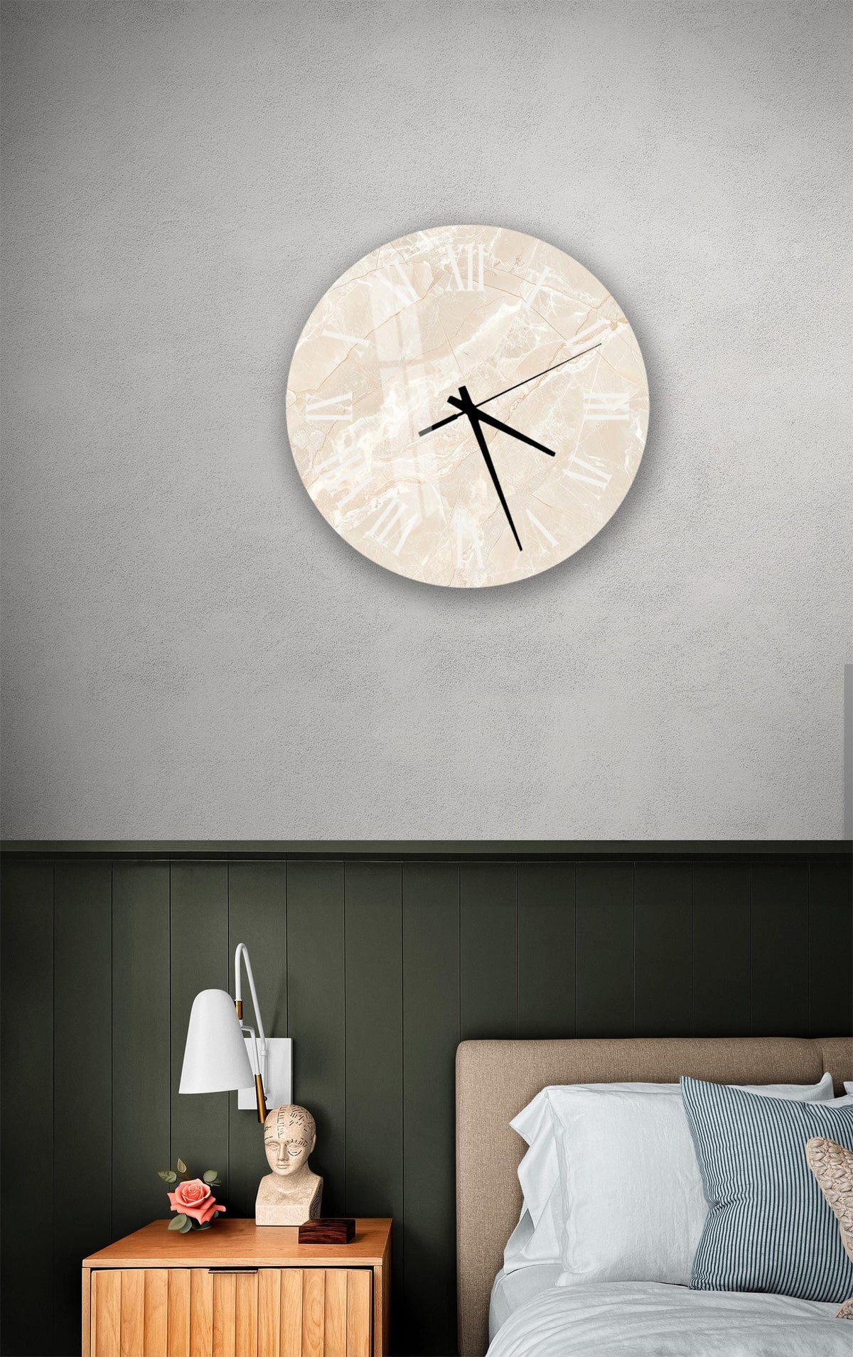 a bedroom setting with a round clock on the wall above a bed. The clock has a marble-like design and is mounted on a gray wall. The bed has a wooden headboard and is covered with white sheets. There is also a nightstand with a lamp and a small statue on it.