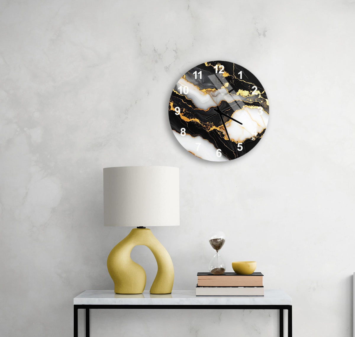 a modern black and white clock with gold accents, mounted on a white wall above a white table with a yellow lamp and a stack of books.