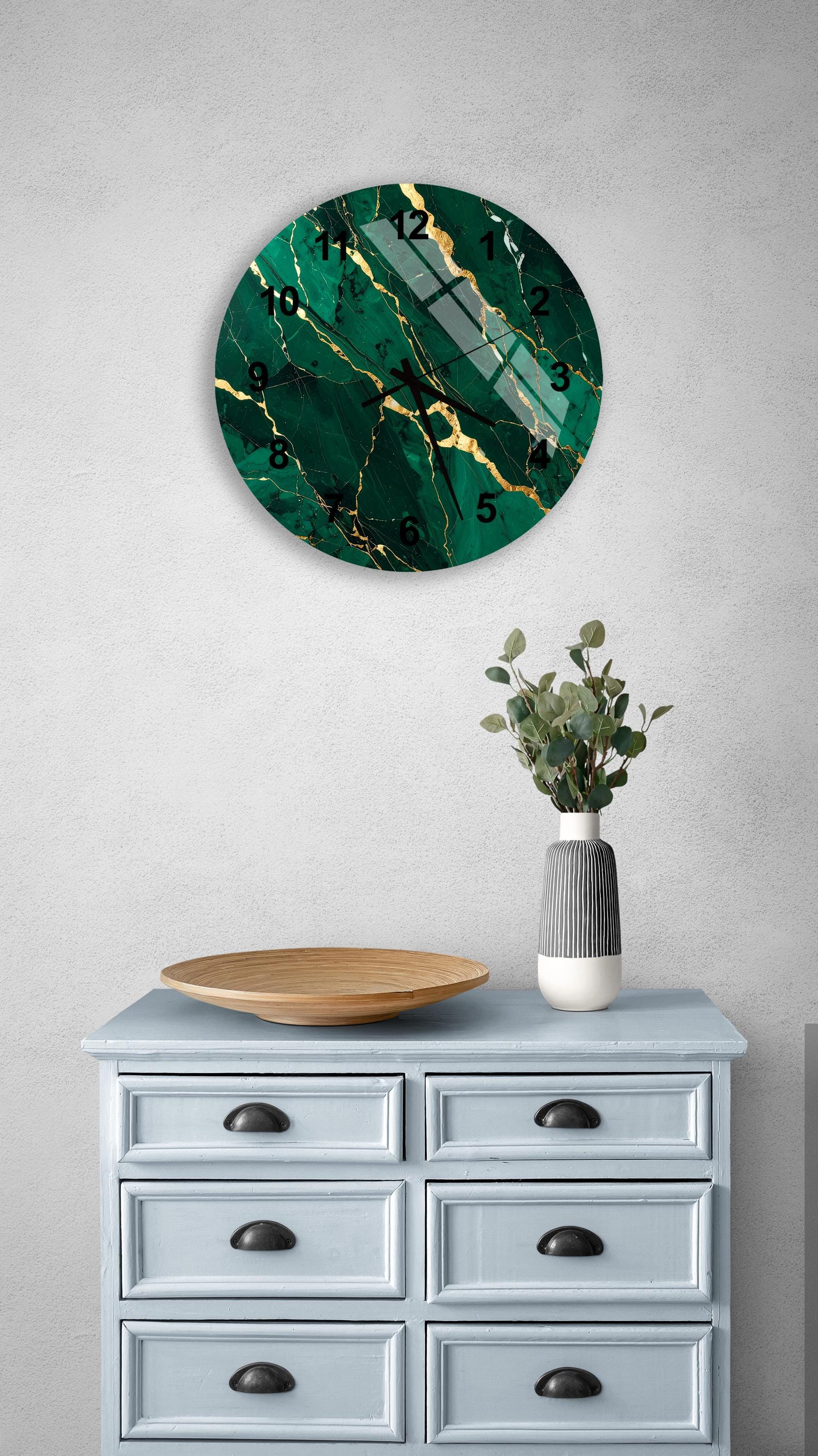 a round clock with a green marble-like design and gold accents, mounted on a wall above a light blue dresser with six drawers. The dresser also has a vase with greenery and a wooden bowl on top.