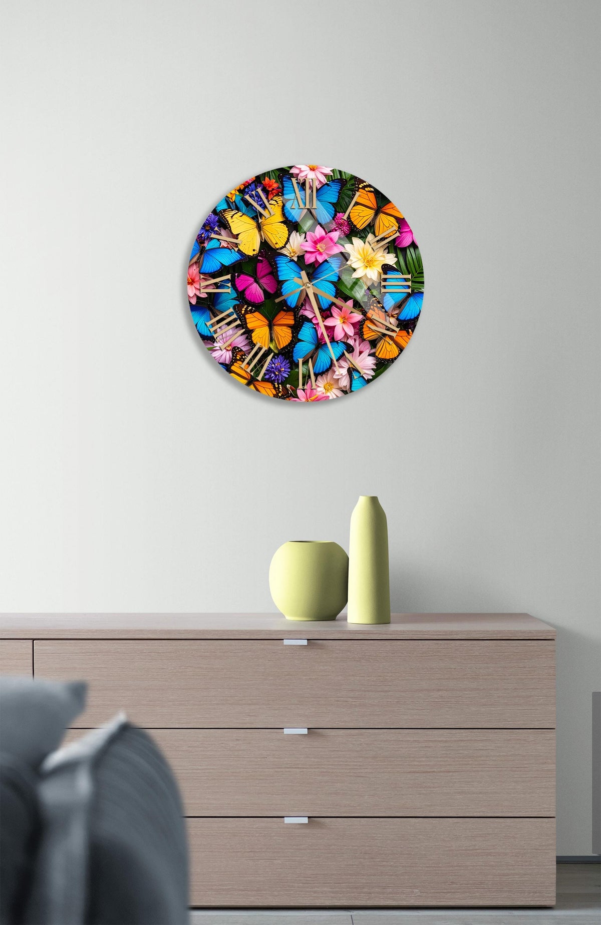 a round clock with a vibrant floral design, mounted on a wall above a wooden dresser with a vase and a small sculpture on top.
