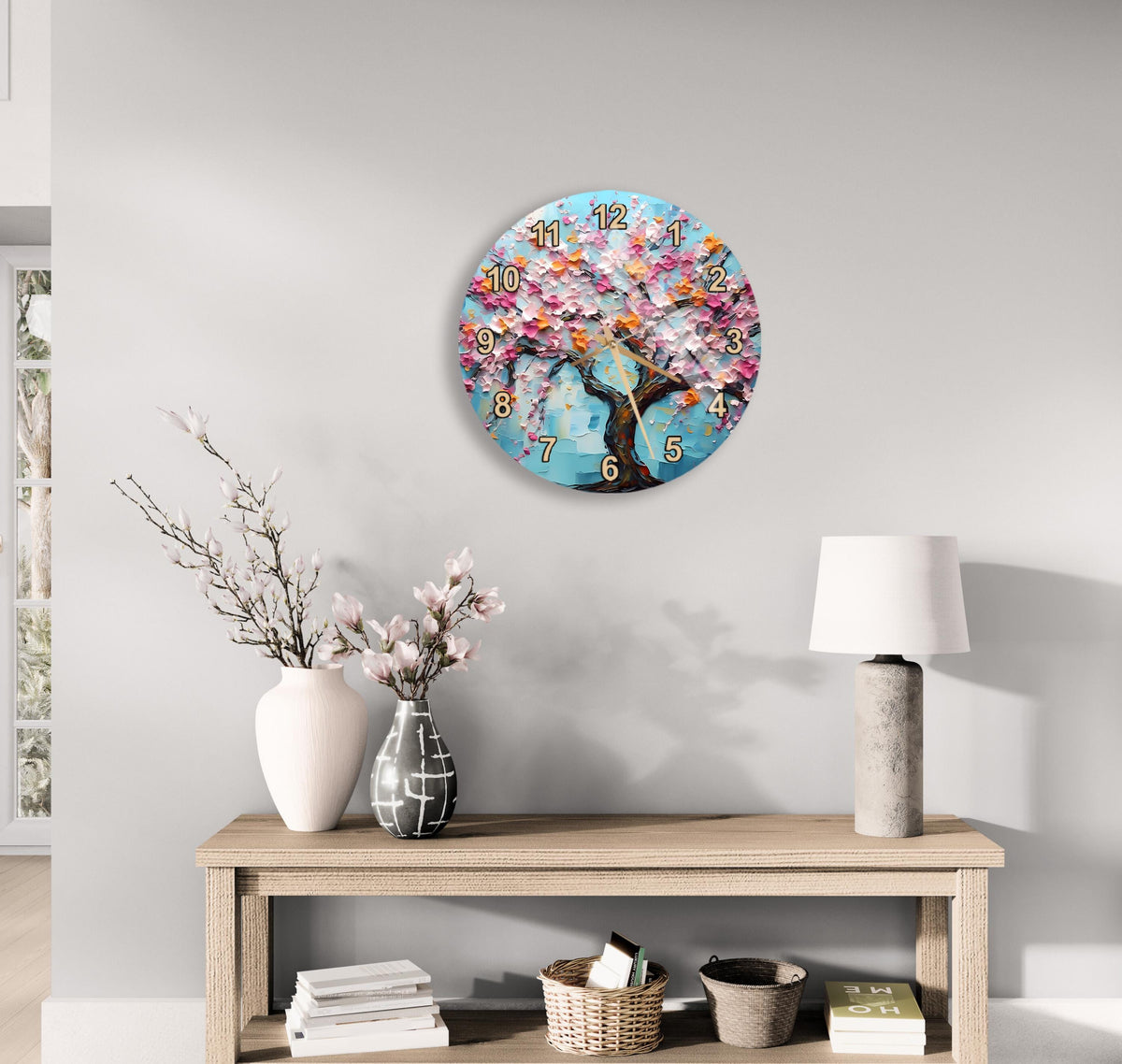 a wall clock with a floral design, a wooden table with a lamp, a vase with pink flowers, and a stack of books on a shelf.