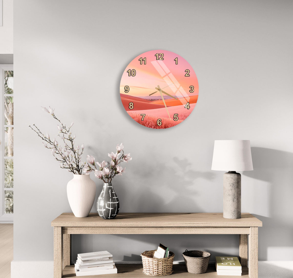 a wall clock with a pink and orange sunset design, a wooden table with a lamp, a vase with flowers, and some books on a shelf.