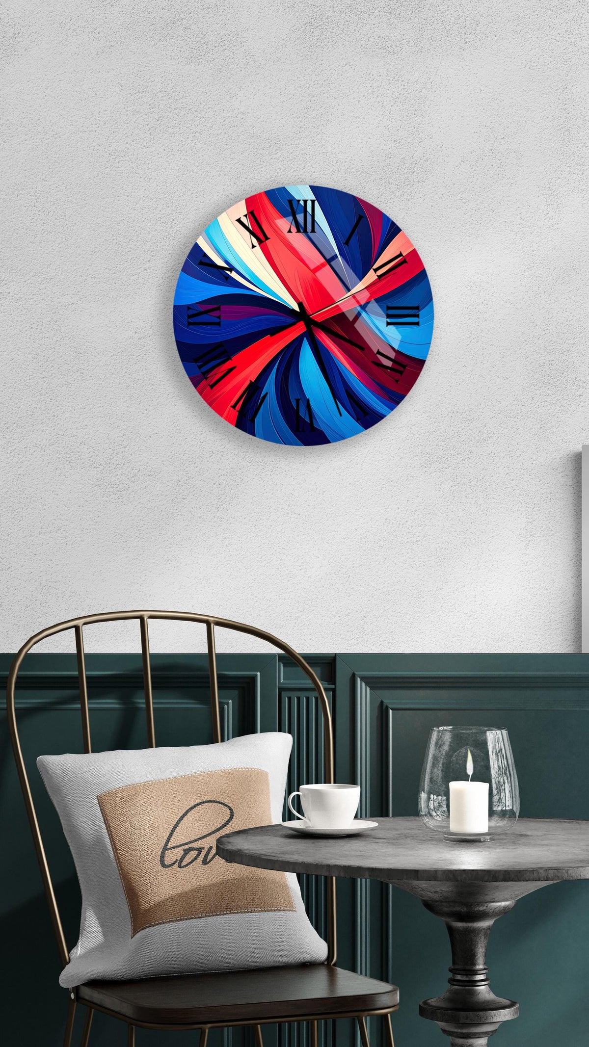 a modern wall clock with a vibrant abstract design in shades of blue, red, and orange. The clock is mounted on a white wall above a dining table with a glass candle and a white coffee cup. A white pillow with the word "love" printed on it is also present on the table.