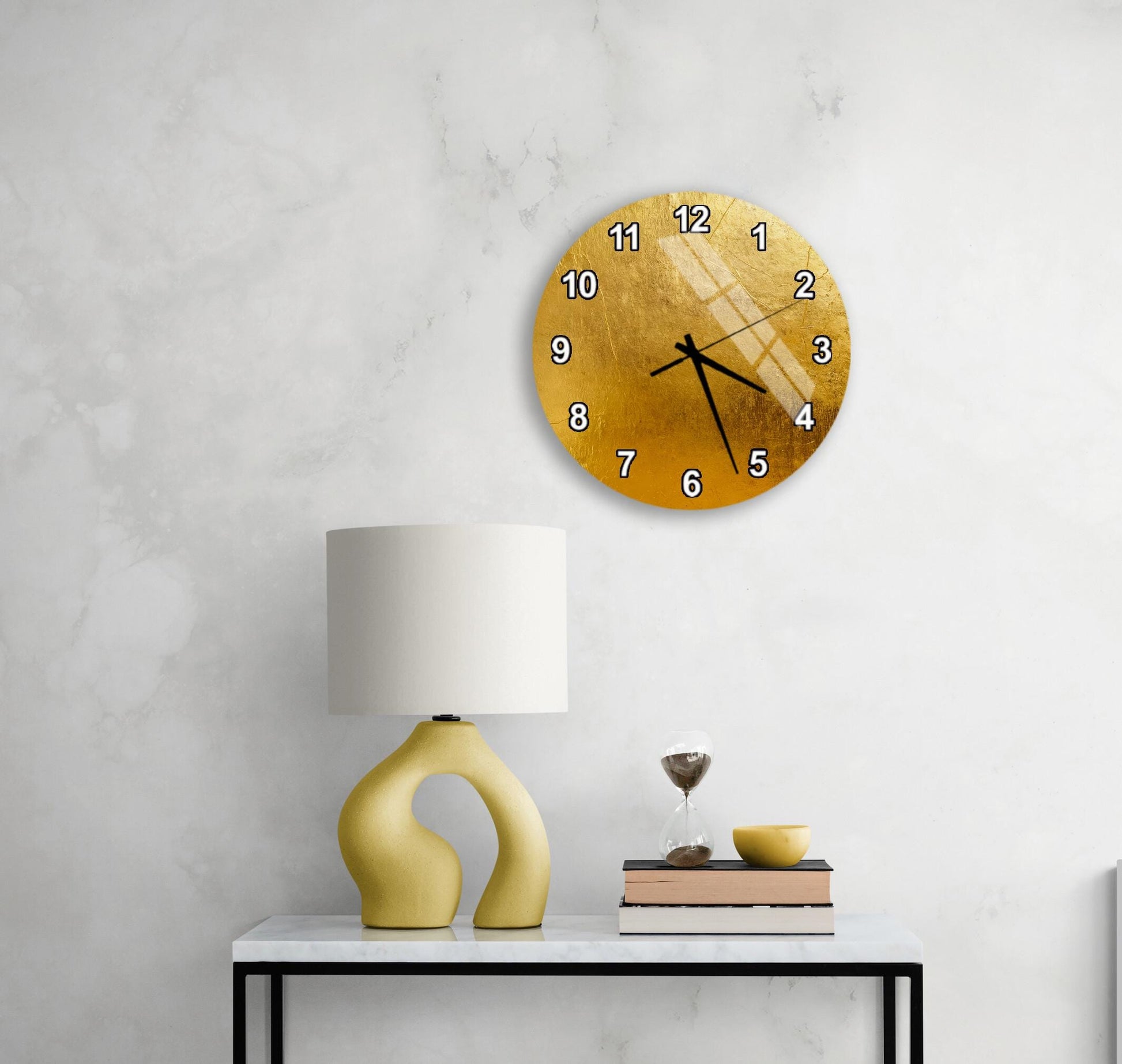 a modern living room with a white marble wall, a round gold clock with a unique design, a yellow lamp with a curved base, a stack of books, and a glass of wine on a white table.