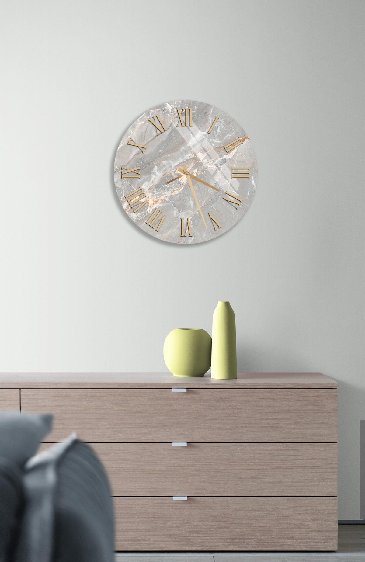 a modern living room with a large, round clock hanging on the wall above a wooden dresser. The clock has a marble-like finish and gold Roman numerals. On the dresser, there are two yellow vases and a small green vase, creating a stylish and cohesive decor.