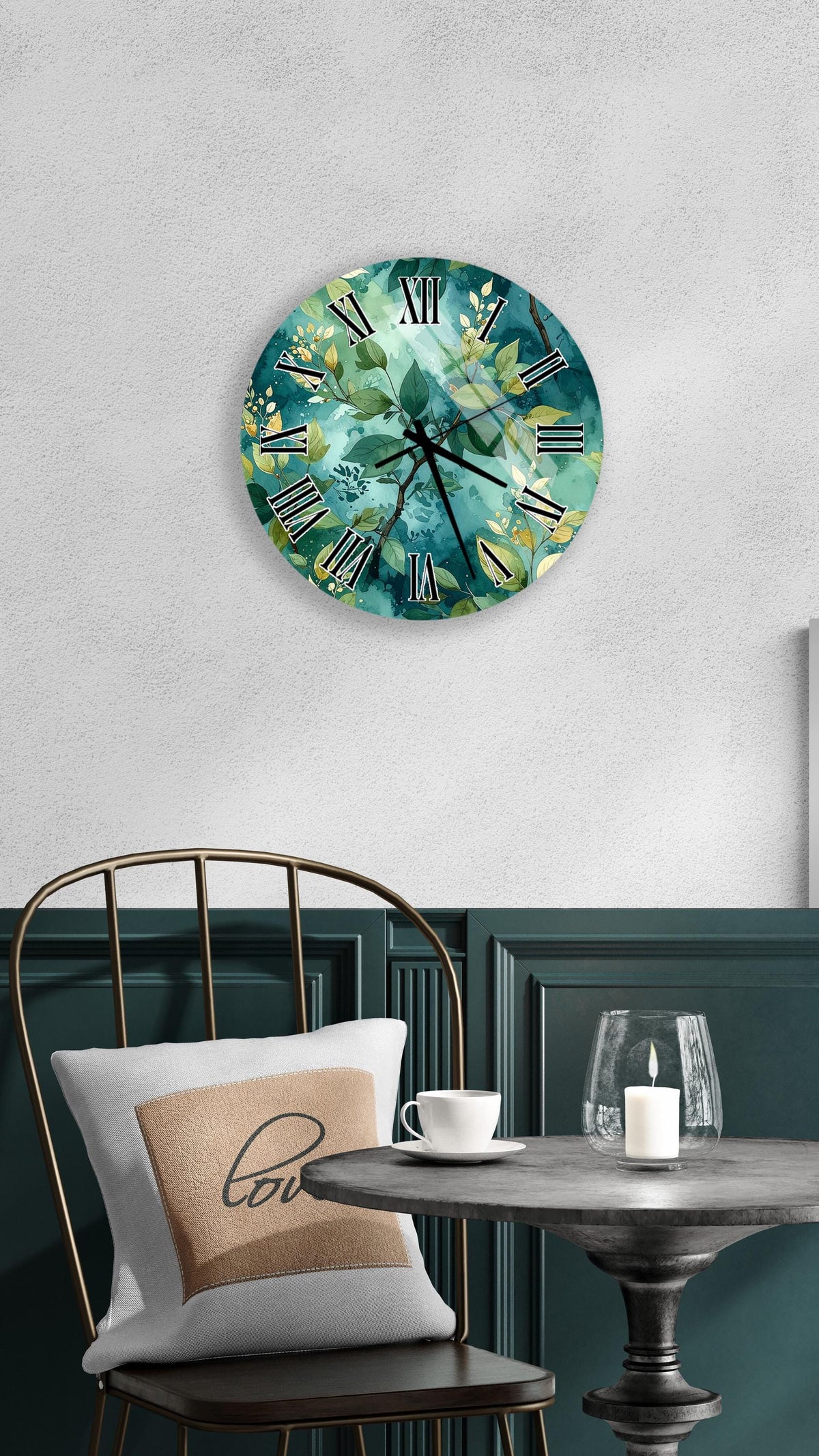 a round wall clock with a green and white floral design, mounted on a white wall above a black metal chair. The chair has a white pillow with the word "love" printed on it. In front of the chair, there is a small table with a glass candle holder and a white teacup.