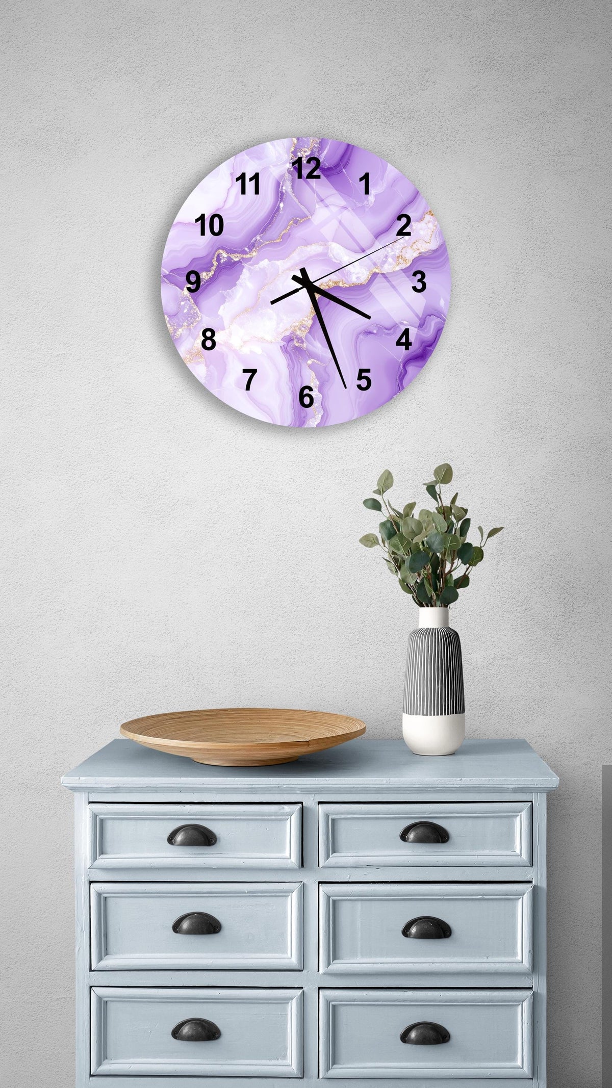 a purple marble wall clock with gold accents, mounted on a wall above a light blue wooden dresser with six drawers. The dresser also has a vase with greenery on it, and there is a wooden bowl on the dresser.