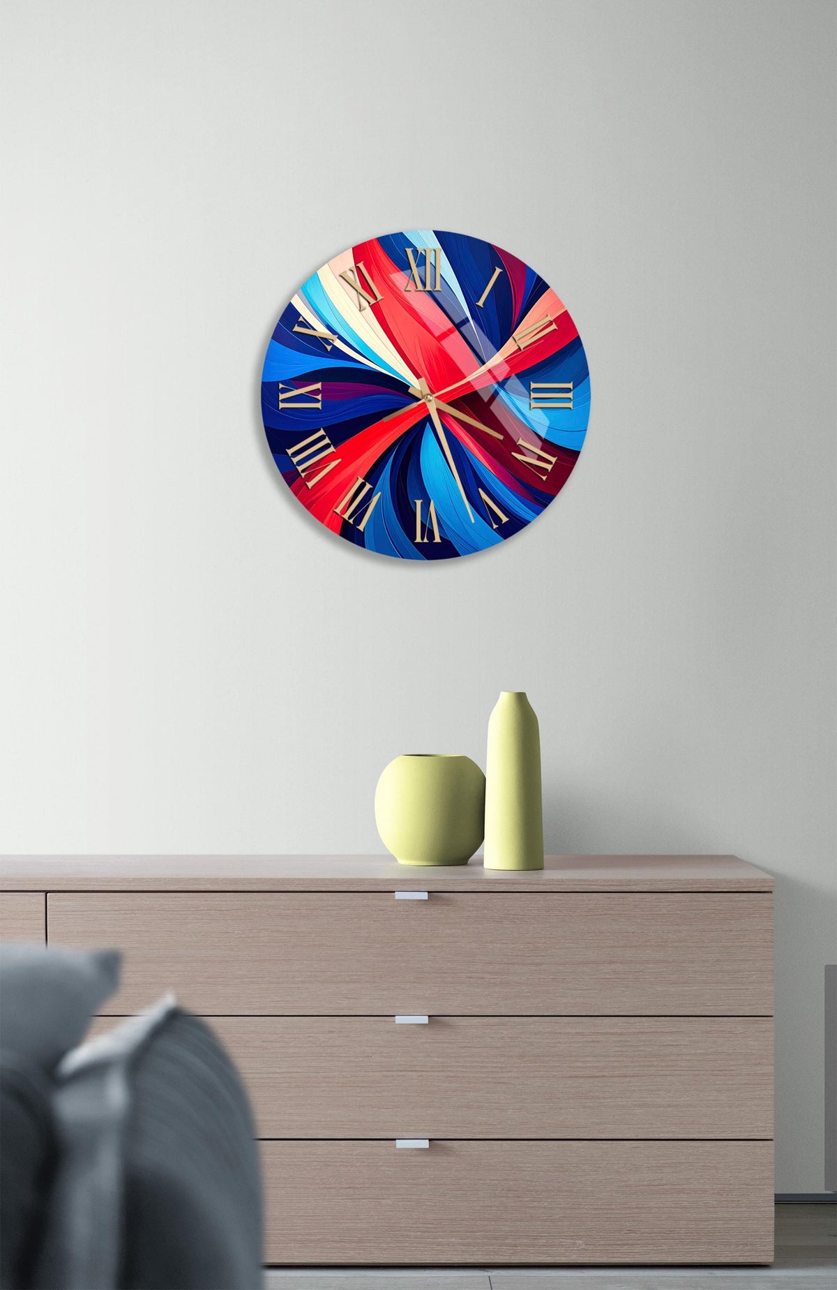 a modern wall clock with a vibrant, abstract design in red, blue, and yellow colors. The clock is mounted on a wall above a wooden dresser with three drawers. The dresser also has a small vase and a yellow pot on it.
