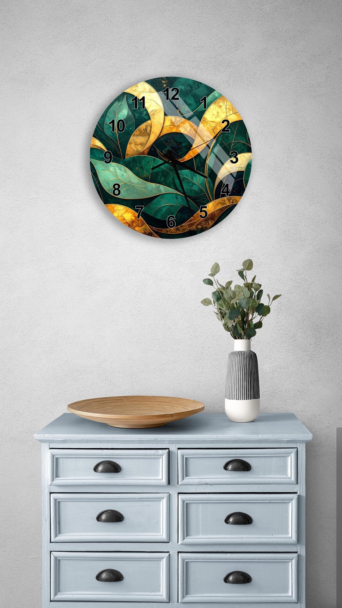 a wall clock with a green and gold leafy design, mounted on a wall above a light blue dresser with six drawers. The dresser also has a wooden plate and a vase with greenery on it.