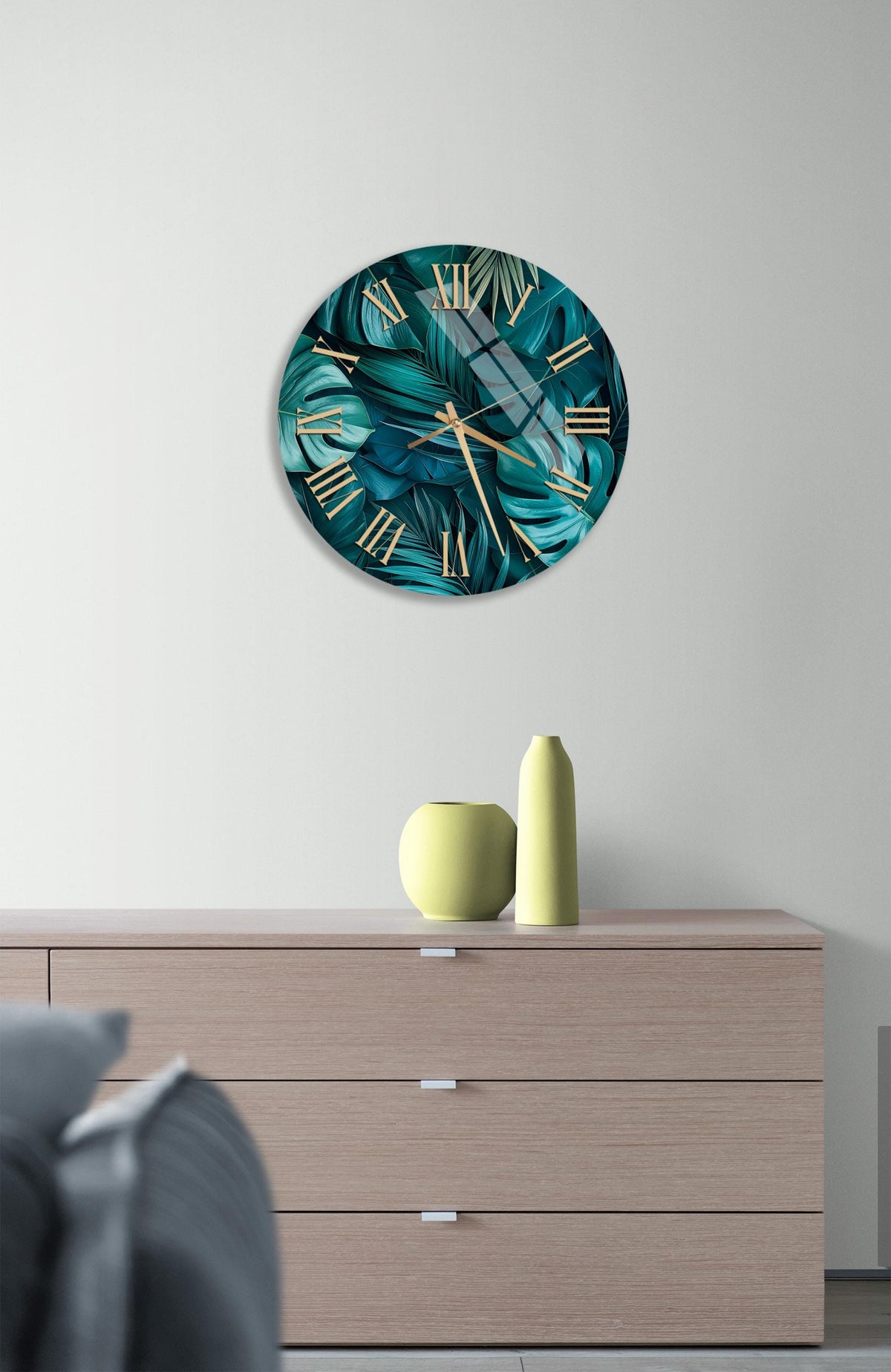 a modern living room with a large wall clock displaying a tropical leaf pattern in shades of green and blue. The clock is mounted above a wooden dresser with three drawers. On the dresser, there are two yellow vases and a small white vase.