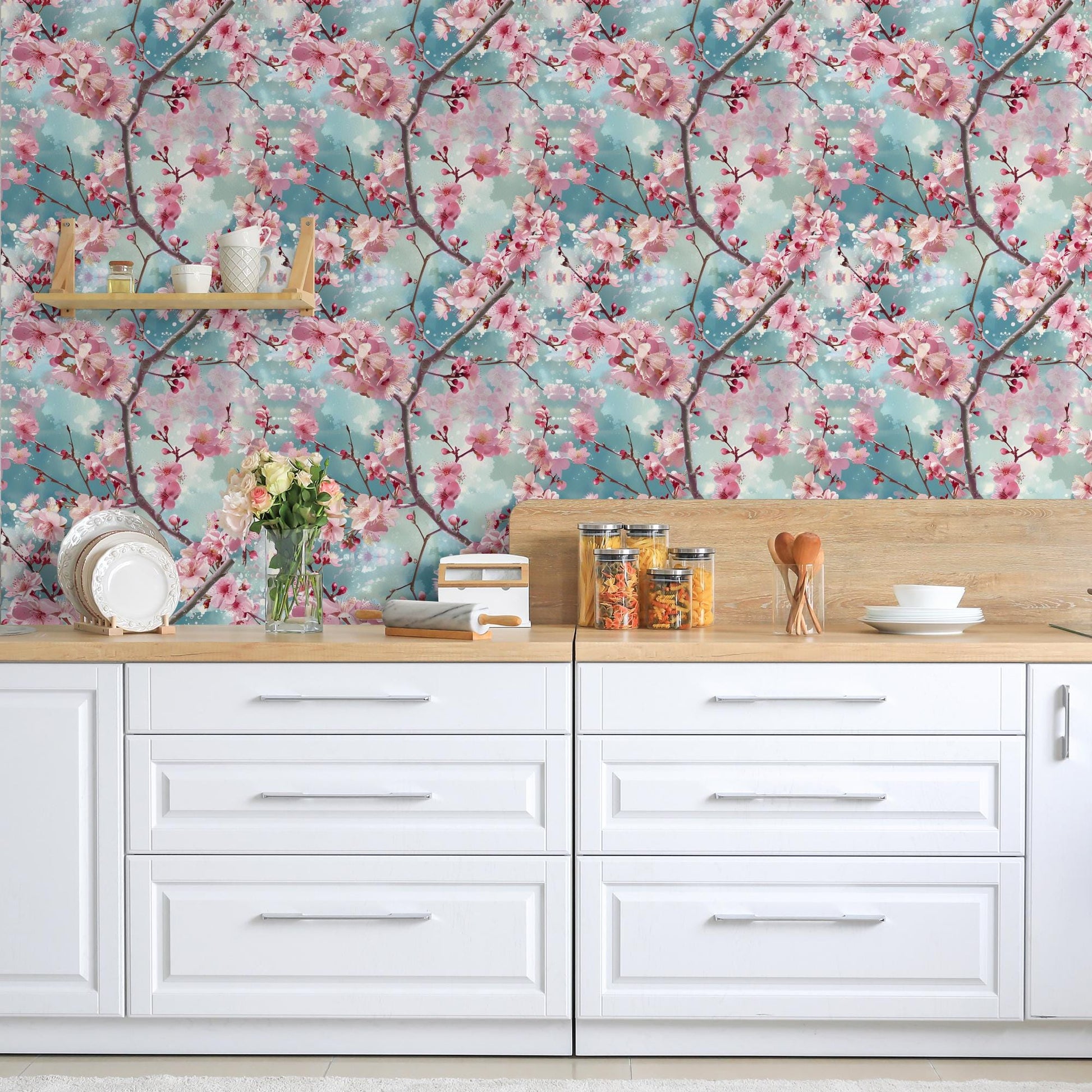 a kitchen counter with white cabinets and drawers, adorned with various kitchen items such as a vase of flowers, a bowl, and a stack of books. The wall behind the counter is decorated with a floral wallpaper, adding a touch of nature and color to the space.