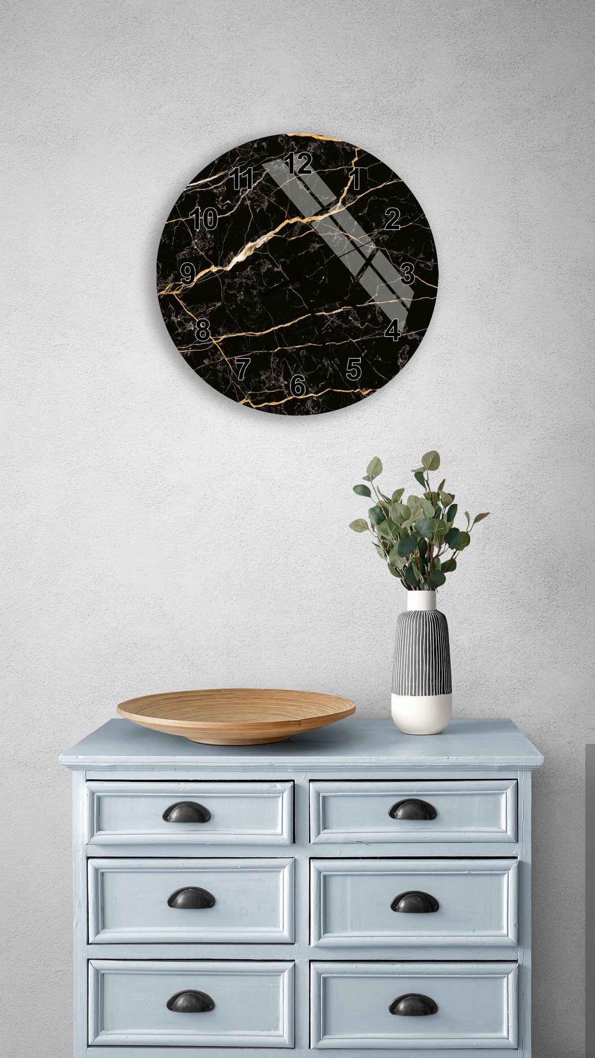 a black marble clock with gold accents, mounted on a wall above a light blue dresser with six drawers. The dresser also has a wooden bowl and a vase with greenery on it.