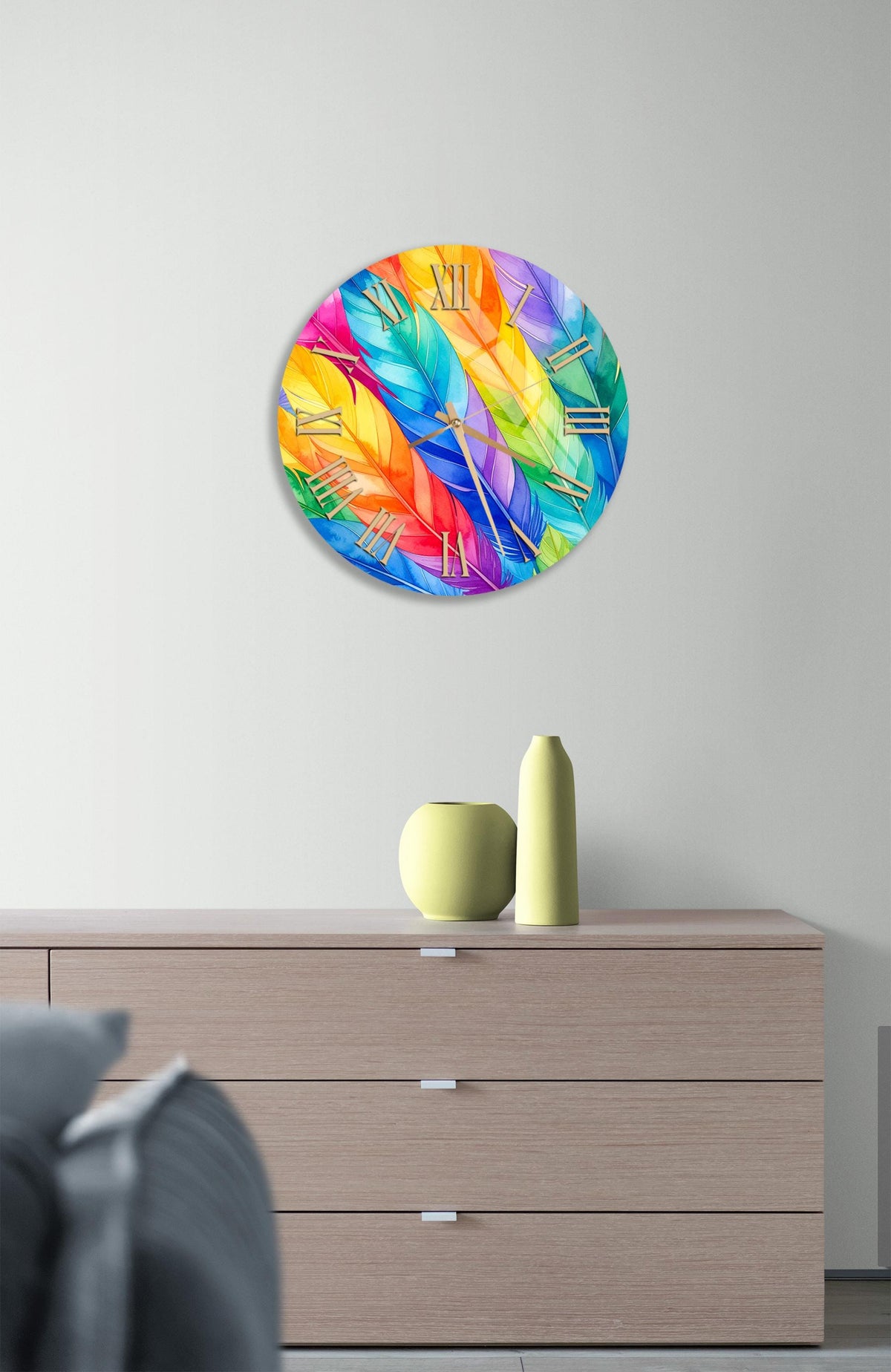 a colorful clock with a vibrant feather design, hanging on a wall above a wooden dresser with a vase and a small sculpture on top.