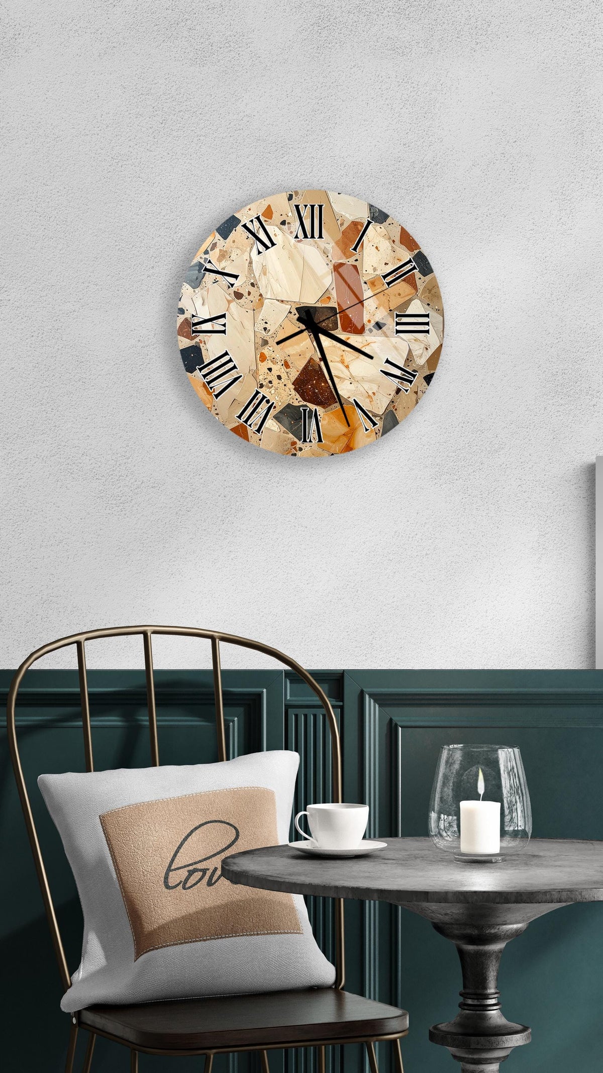 a round wall clock with a marble-like design, mounted on a wall above a table with a white tablecloth. The table has a glass candle and a white coffee cup on it. A white pillow with the word "love" printed on it is also present on the table.