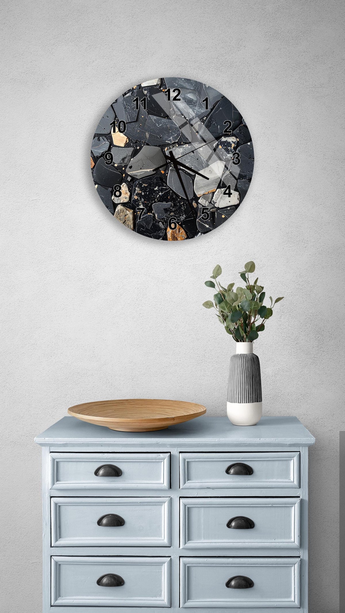 a wall clock with a black and white design, mounted on a wall above a light blue dresser with six drawers. The dresser also has a wooden bowl and a vase with greenery on it.