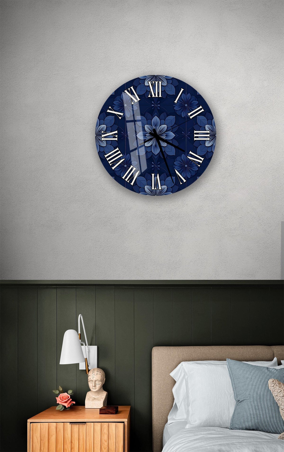 a bedroom setting with a blue floral wall clock hanging above a bed. The clock has a white face with black Roman numerals and a blue floral design. The room has a green wall, a wooden nightstand with a lamp, and a bed with white sheets and pillows.