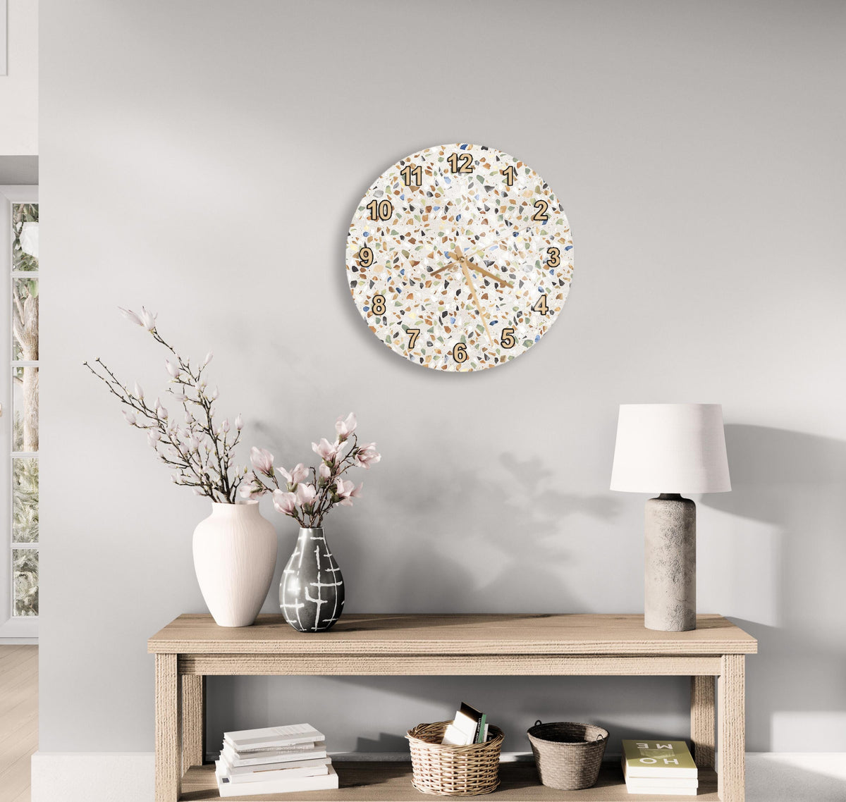 a modern and minimalist living room with a wooden console table, a round wall clock with a speckled design, a vase with pink flowers, and a lamp on a side table.