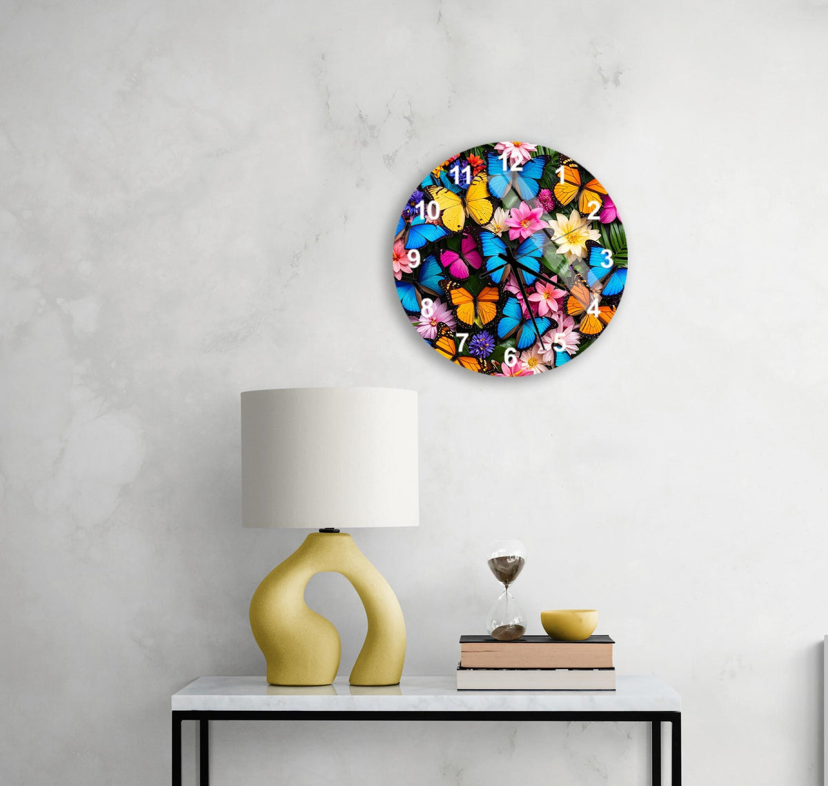 a wall clock with a butterfly design, a yellow table lamp, a glass of water, and a stack of books on a white table.