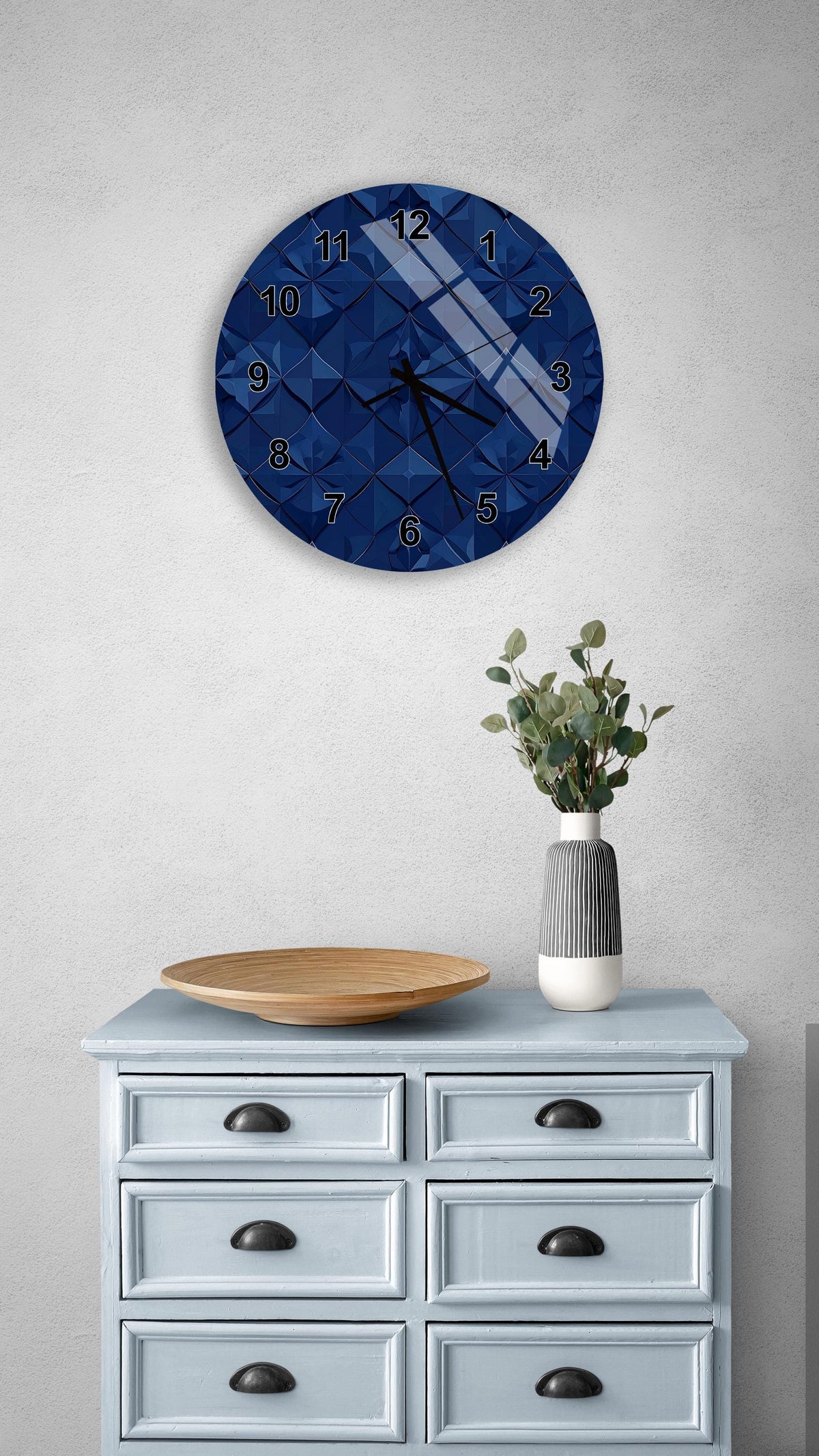 a blue clock with a geometric pattern on the wall above a light blue dresser with six drawers. The dresser also has a wooden bowl and a vase with greenery on it.