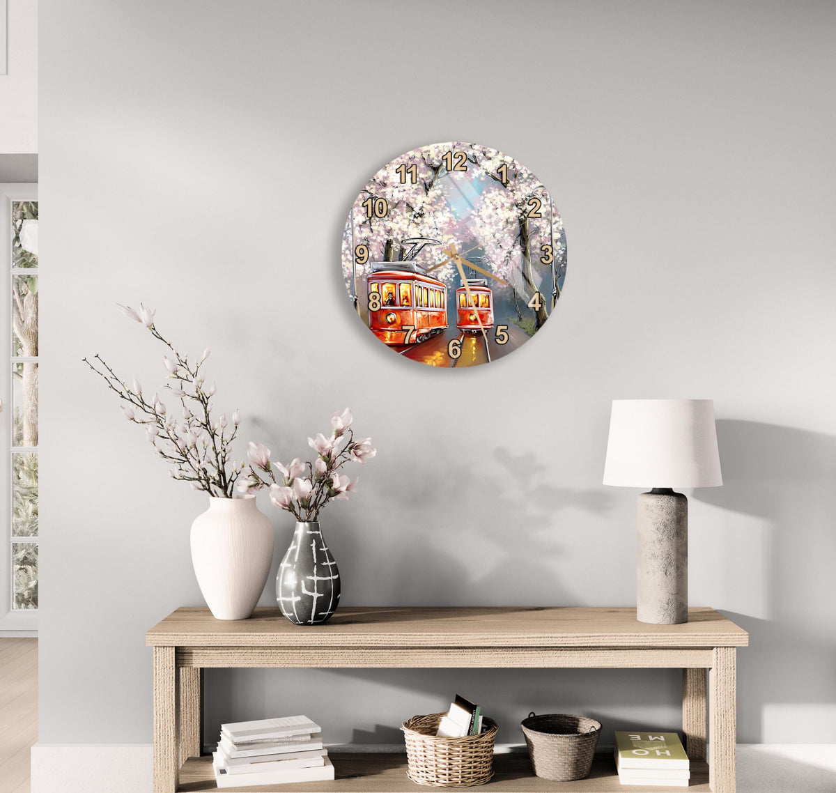 a round clock with a floral design hanging on a wall above a wooden table with various decorative items, including a vase with flowers, a lamp, and a stack of books.